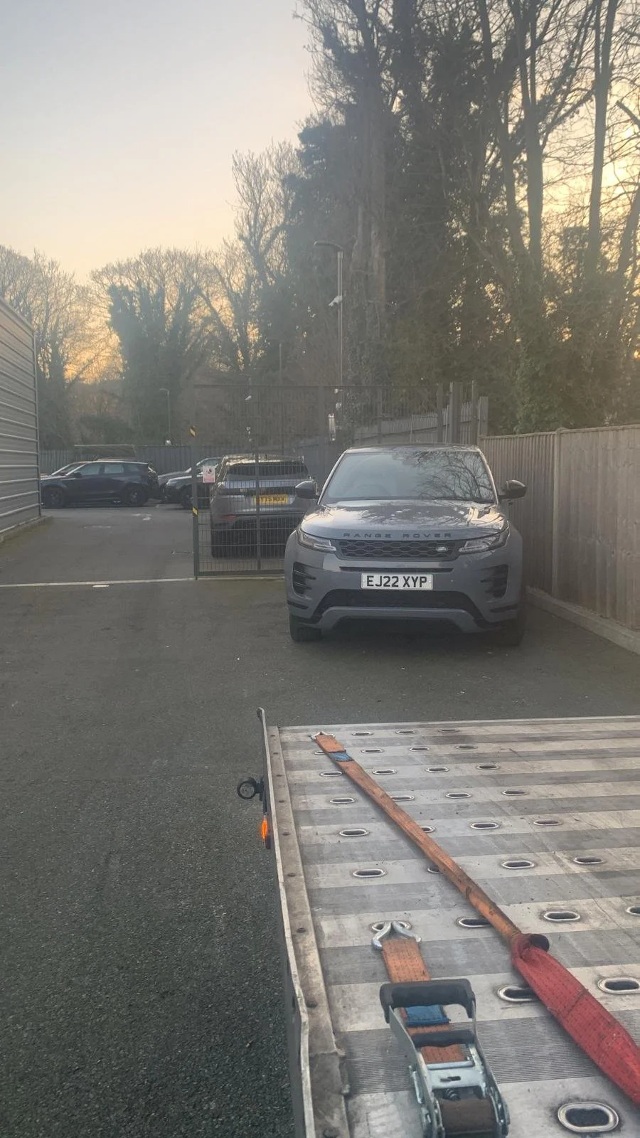 A parking lot with a Range Rover parked near a wooden fence, with other cars visible in the background. There is also a flatbed trailer with a red strap and a tie-down winch in the foreground.