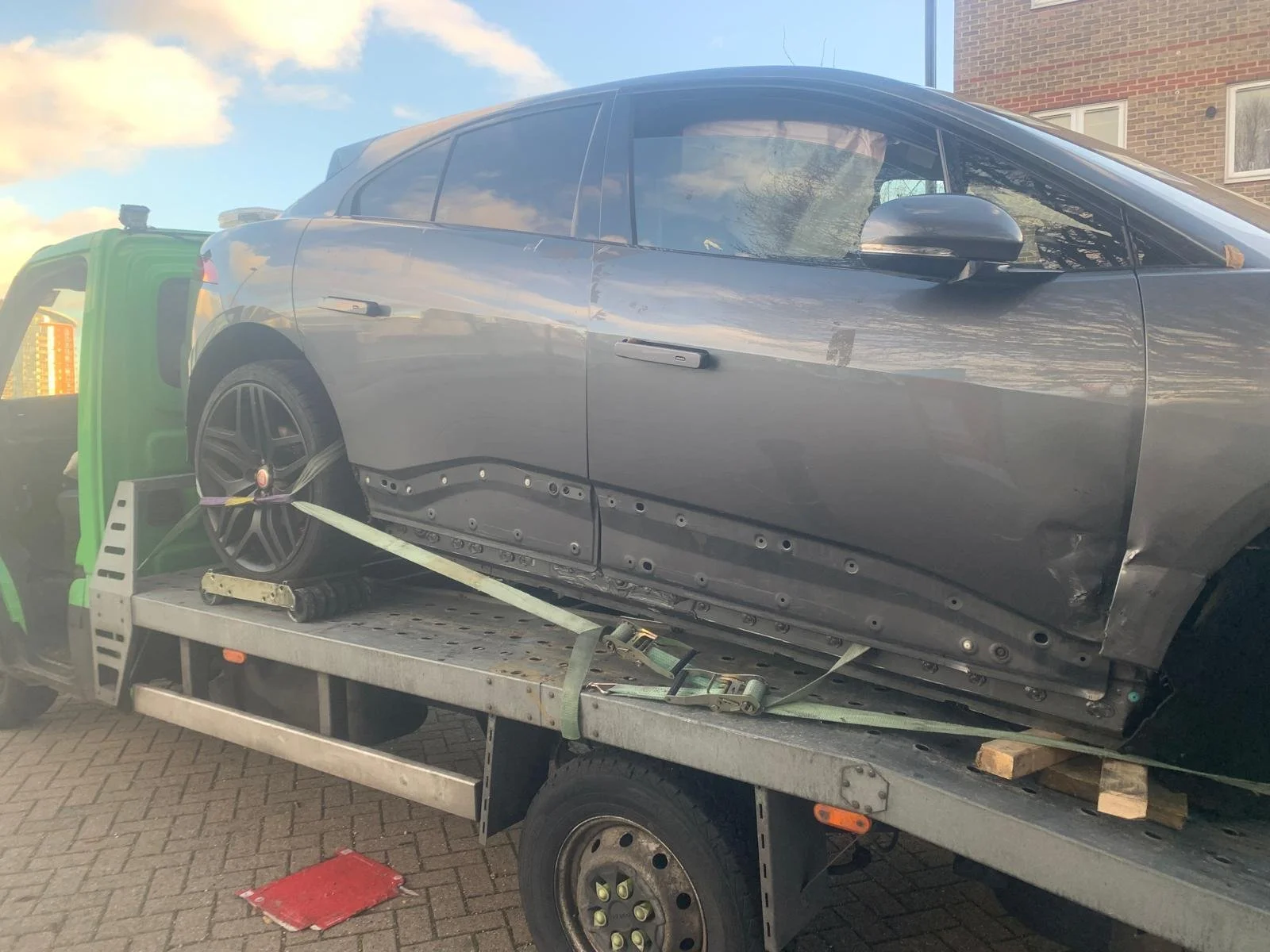 A grey Tesla Model S with visible damages on the side, loaded onto a flatbed tow truck. The side panel and door are dented and scratched, and it is secured with straps for transport.