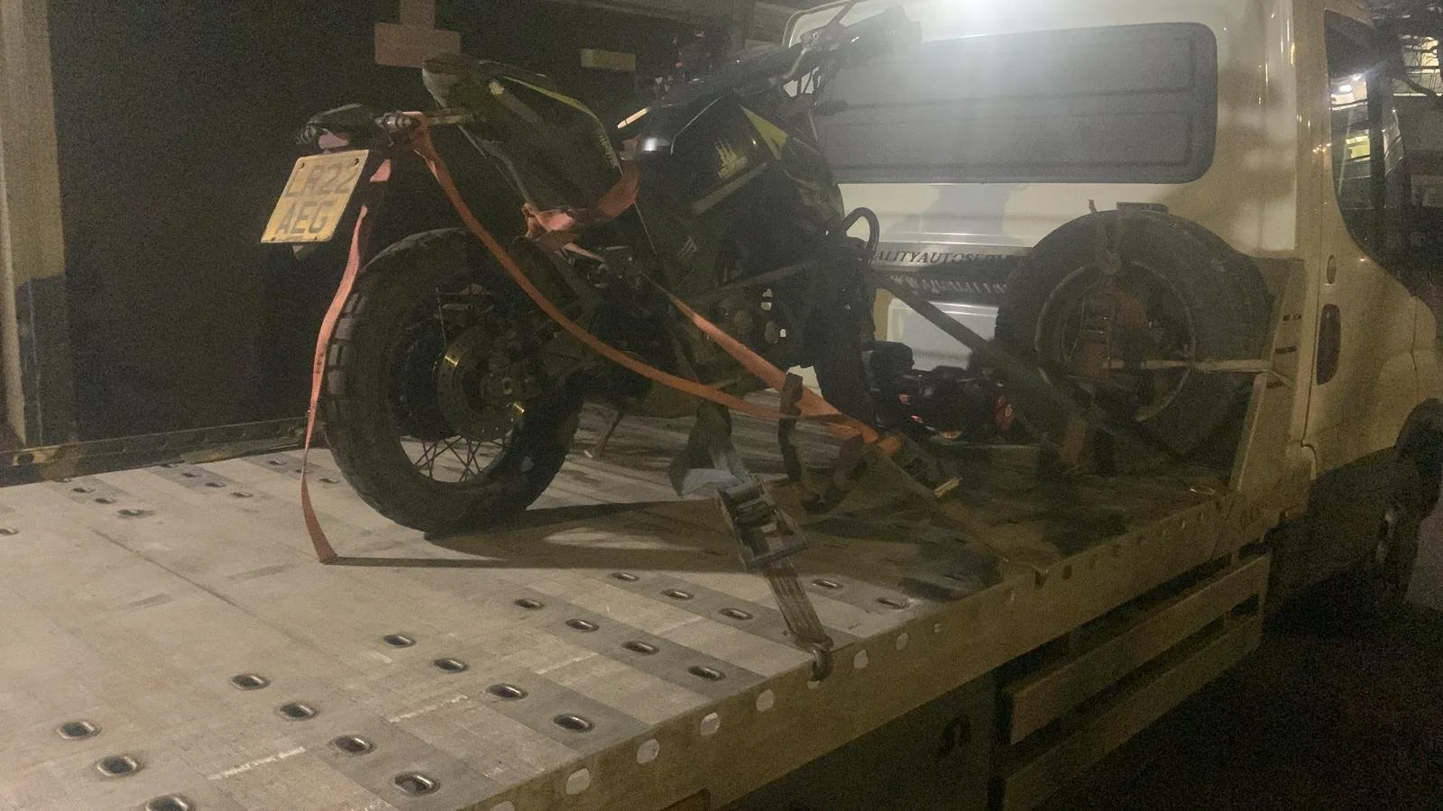 A motorcycle secured with orange straps on a flatbed truck, with another vehicle in the background.