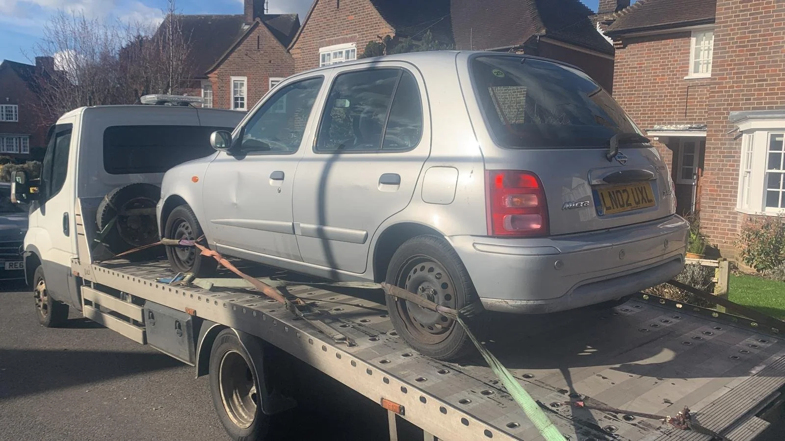 A silver compact car loaded onto a flatbed tow truck, secured with straps, parked in a residential neighborhood with brick houses and a blue sky.