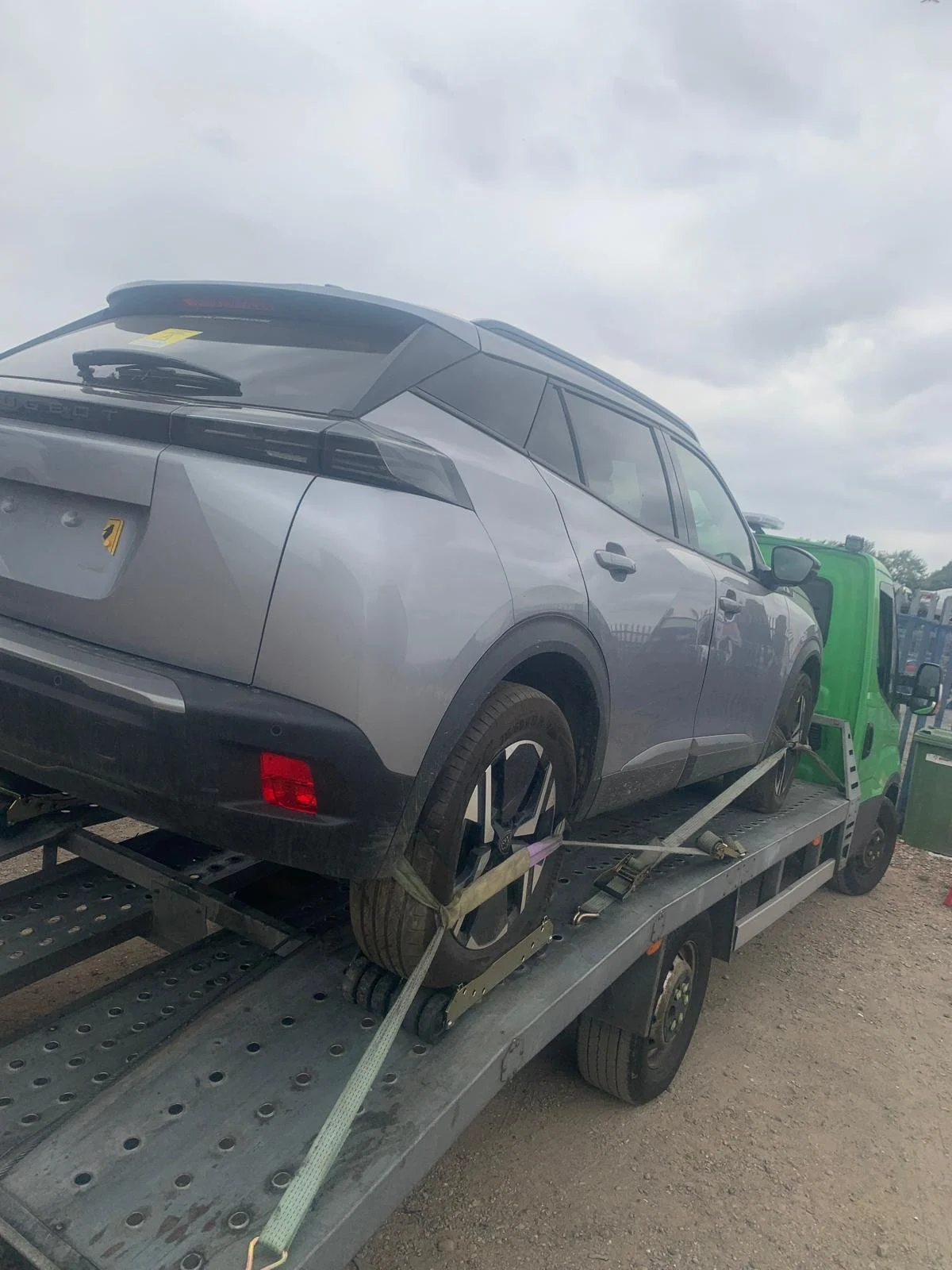 A silver Land Rover SUV on a flatbed tow truck, secured with straps, during daytime with overcast skies.