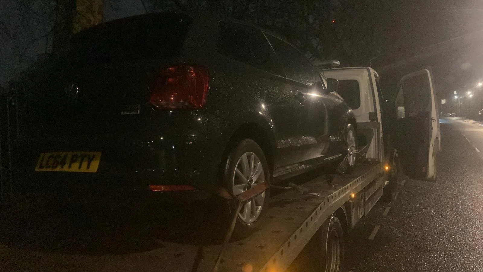 A black sedan on a flatbed tow truck at night, with its driver side door open on a wet road.