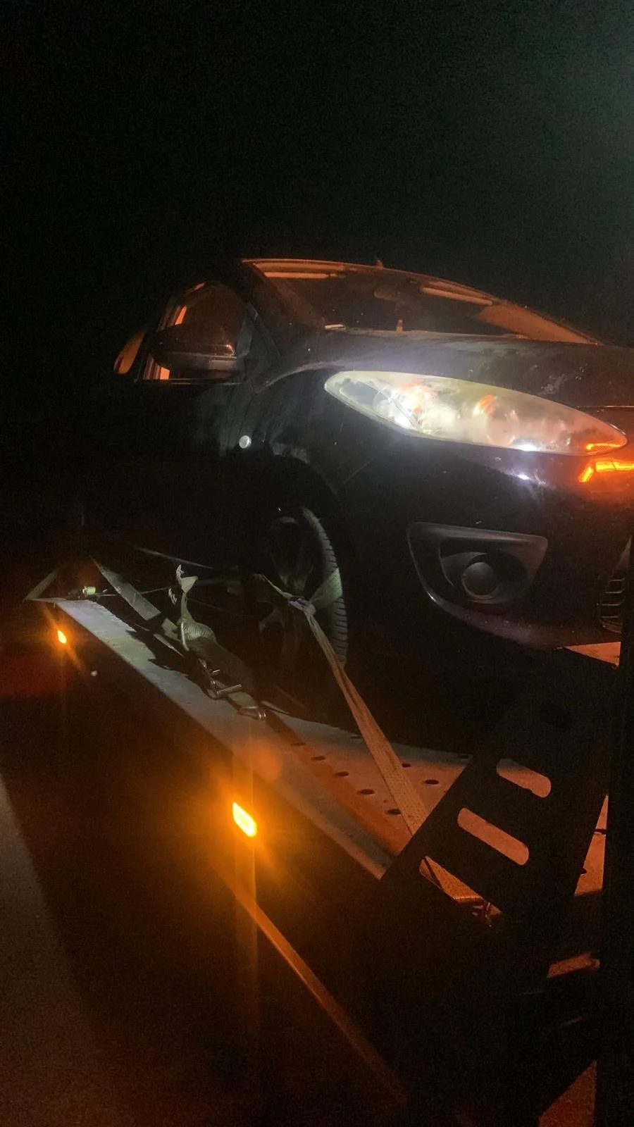 A black car is being loaded onto a flatbed tow truck at night, with the car's headlights illuminated in darkness.