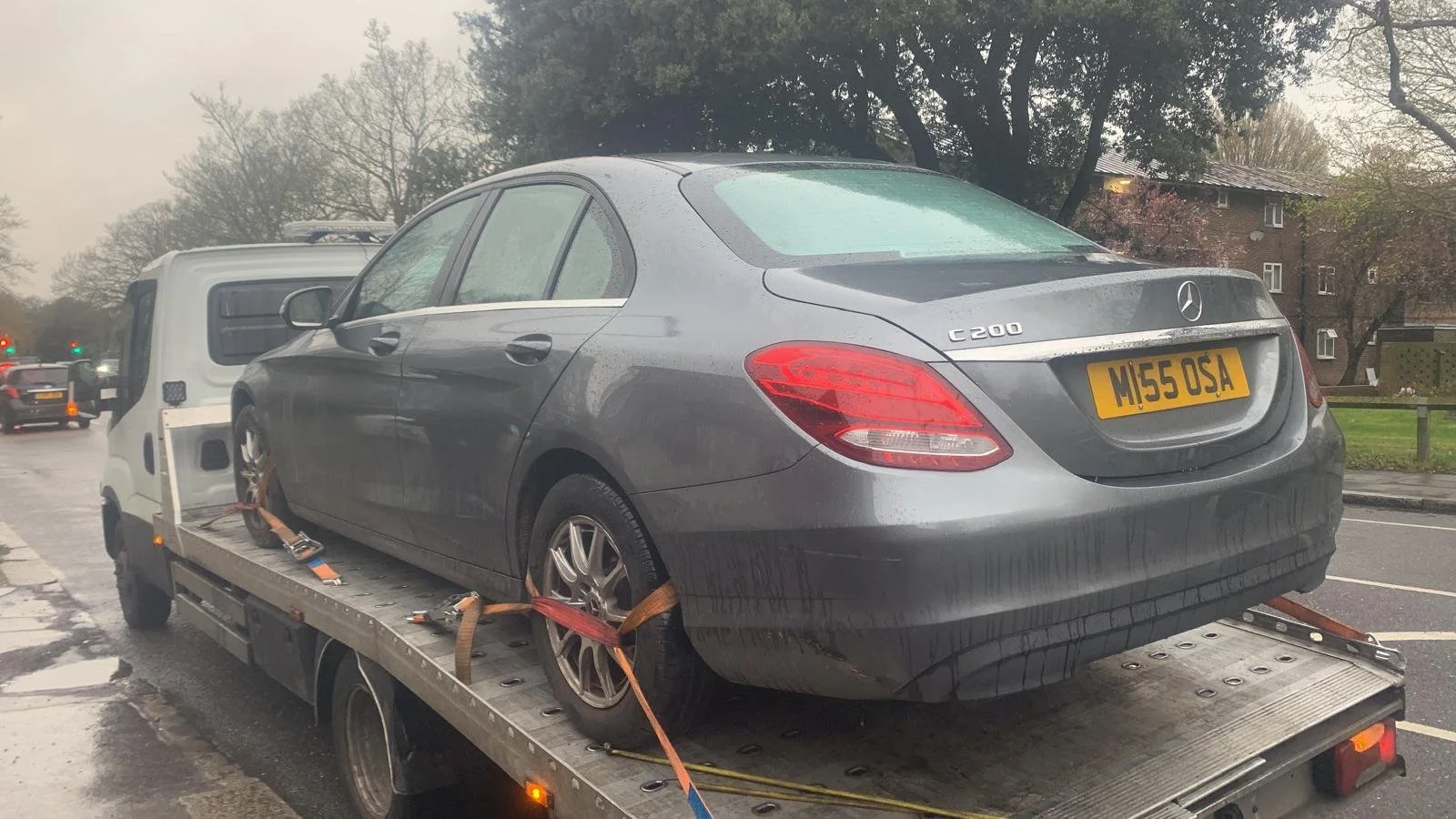 A gray Mercedes-Benz C200 is loaded onto a flatbed tow truck, secured with straps, on a rainy day in a residential or urban area.
