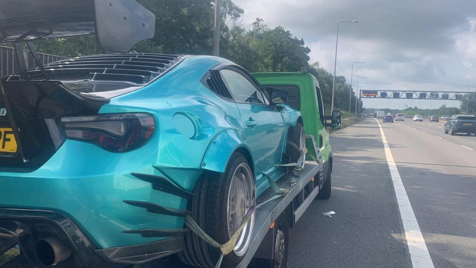 A blue sports car is on a flatbed tow truck on the side of a highway, with multiple lanes of traffic in the distance under a partly cloudy sky.