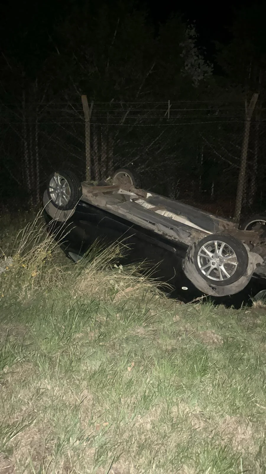 A black car flipped upside down in a grassy area at night, with a chain link fence and trees in the background.