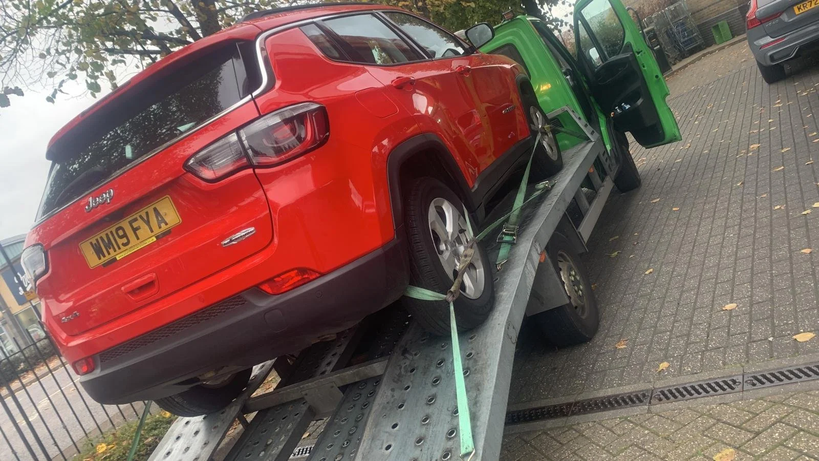 A red Jeep is secured with green straps on a flatbed tow truck, parked on a brick sidewalk with other vehicles nearby.