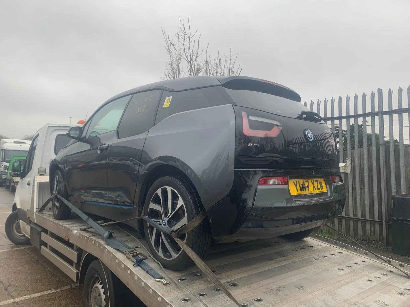 A black BMW i3 electric car is secured on a flatbed tow truck, with a fence and parked vehicles visible in the background.