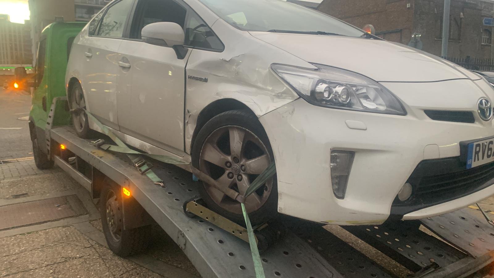 A white Toyota hybrid car on a flatbed tow truck, with noticeable damage to the front right side including a dented bumper and fender.