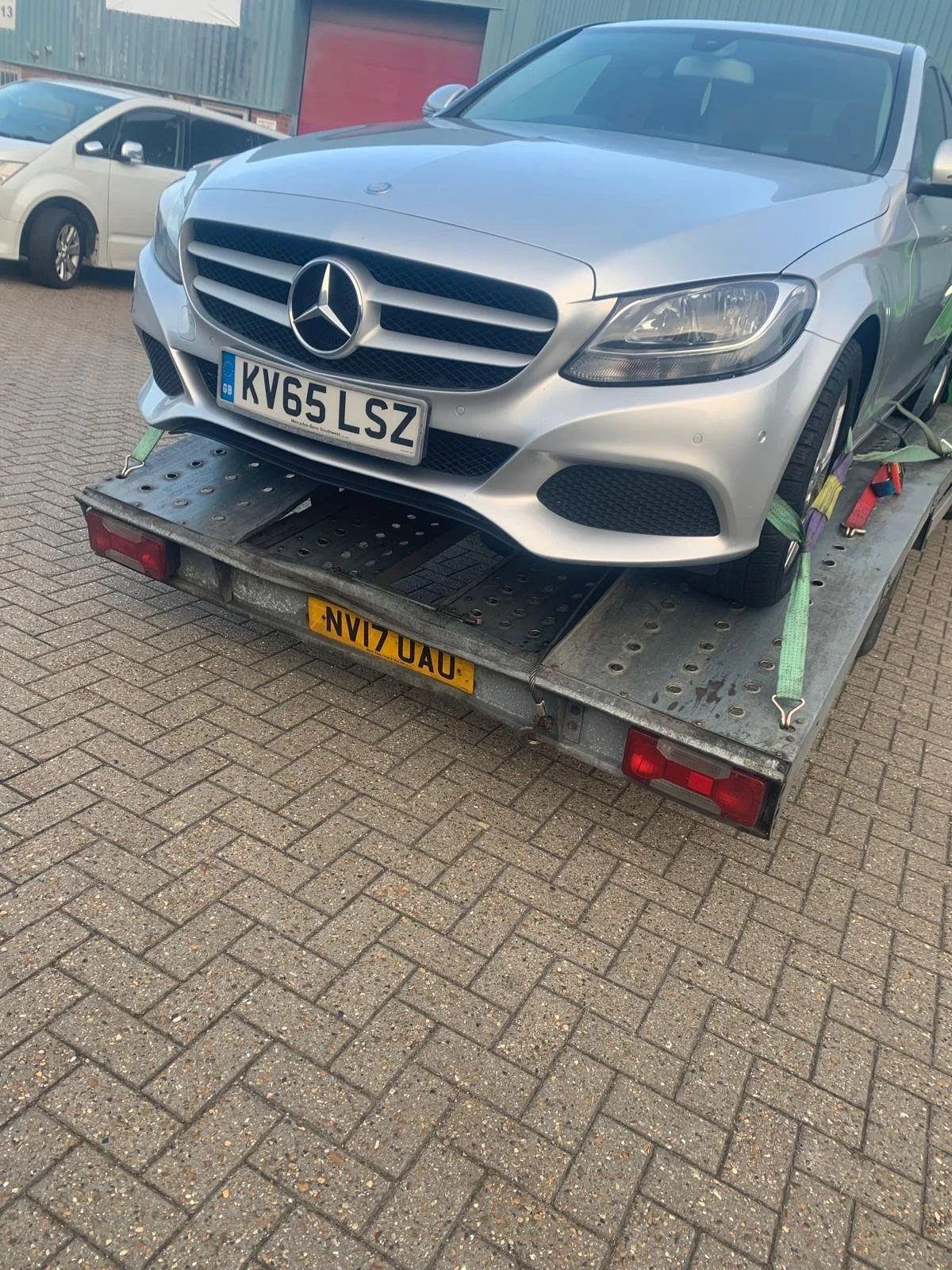 Silver Mercedes-Benz car loaded on a flatbed tow truck, secured with green, purple, and red straps, parked on brick pavement with other vehicles nearby.