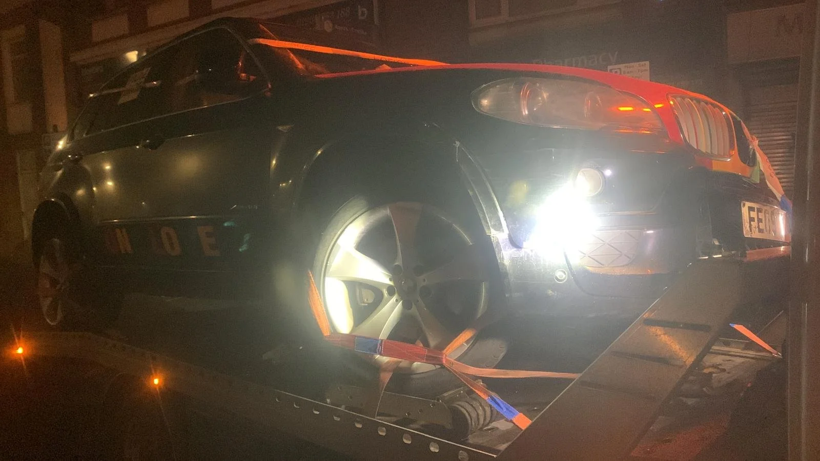 A black car with a visible British license plate on a tow truck at night, illuminated by bright lights.