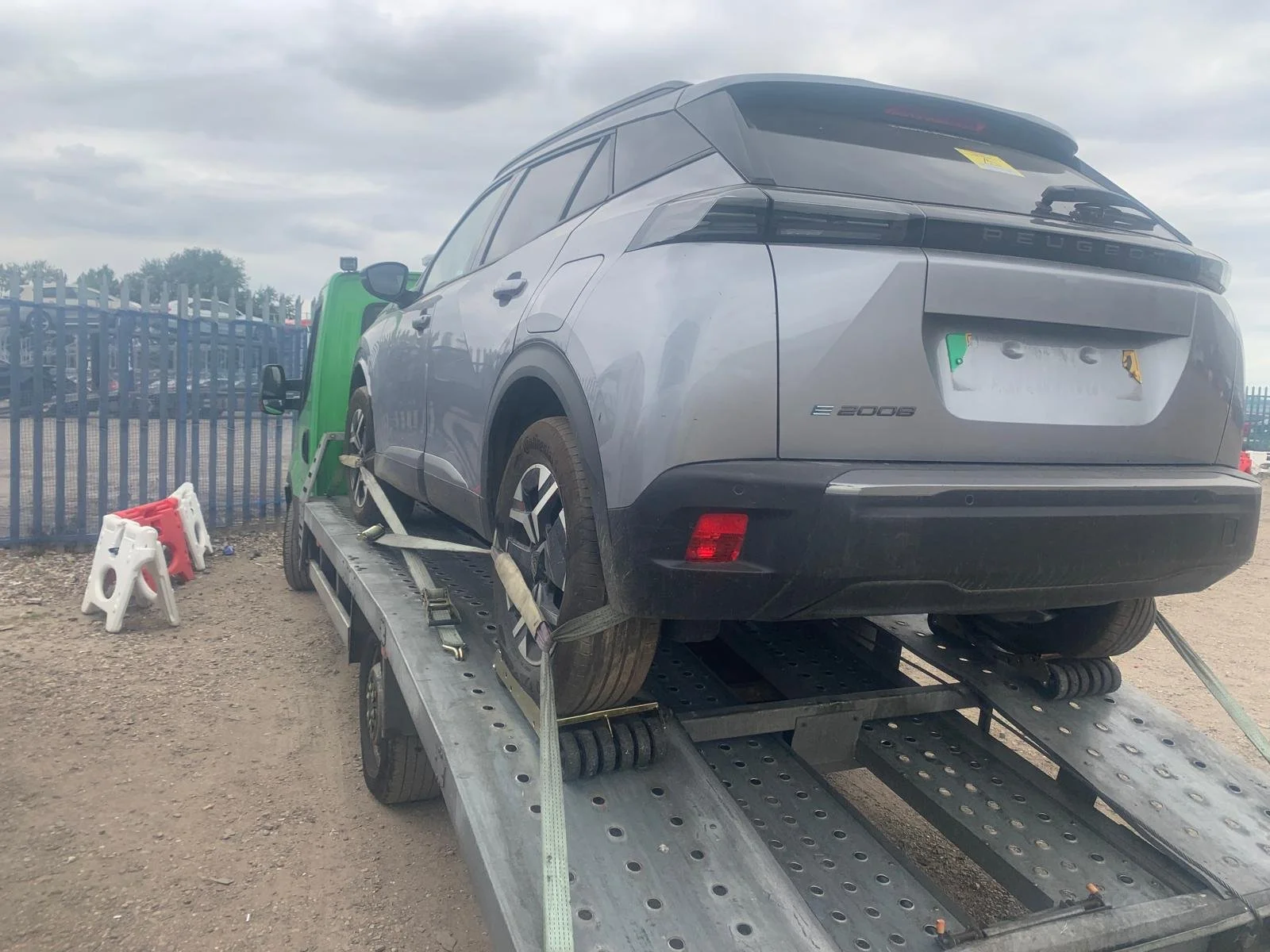 A gray Peugeot E-2008 compact electric SUV on a flatbed tow truck, secured with straps, with a fenced area and parking lot in the background under a cloudy sky.