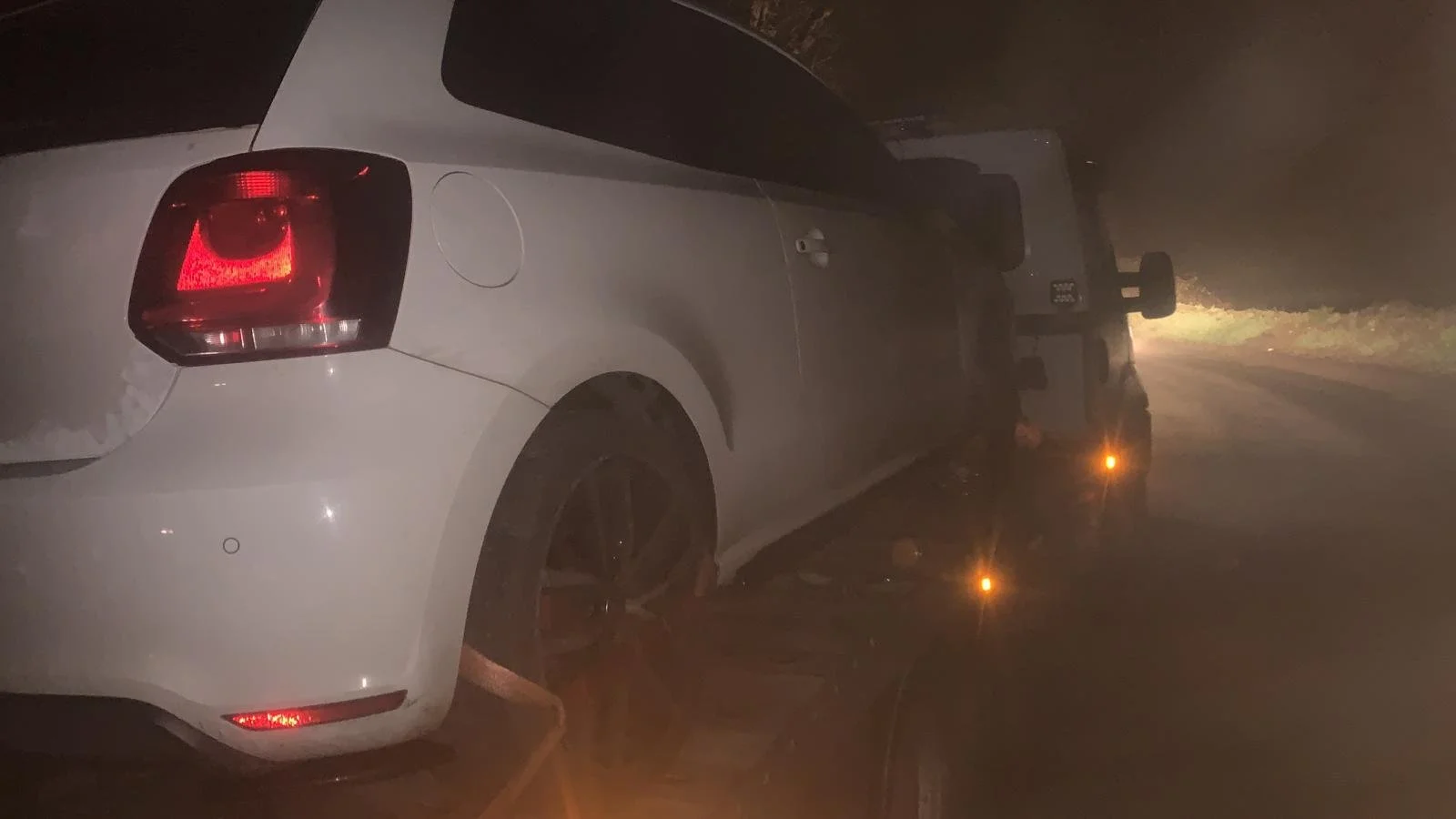 Rear view of a white sedan car on a dark, foggy road at night with an emergency vehicle with flashing lights behind it.
