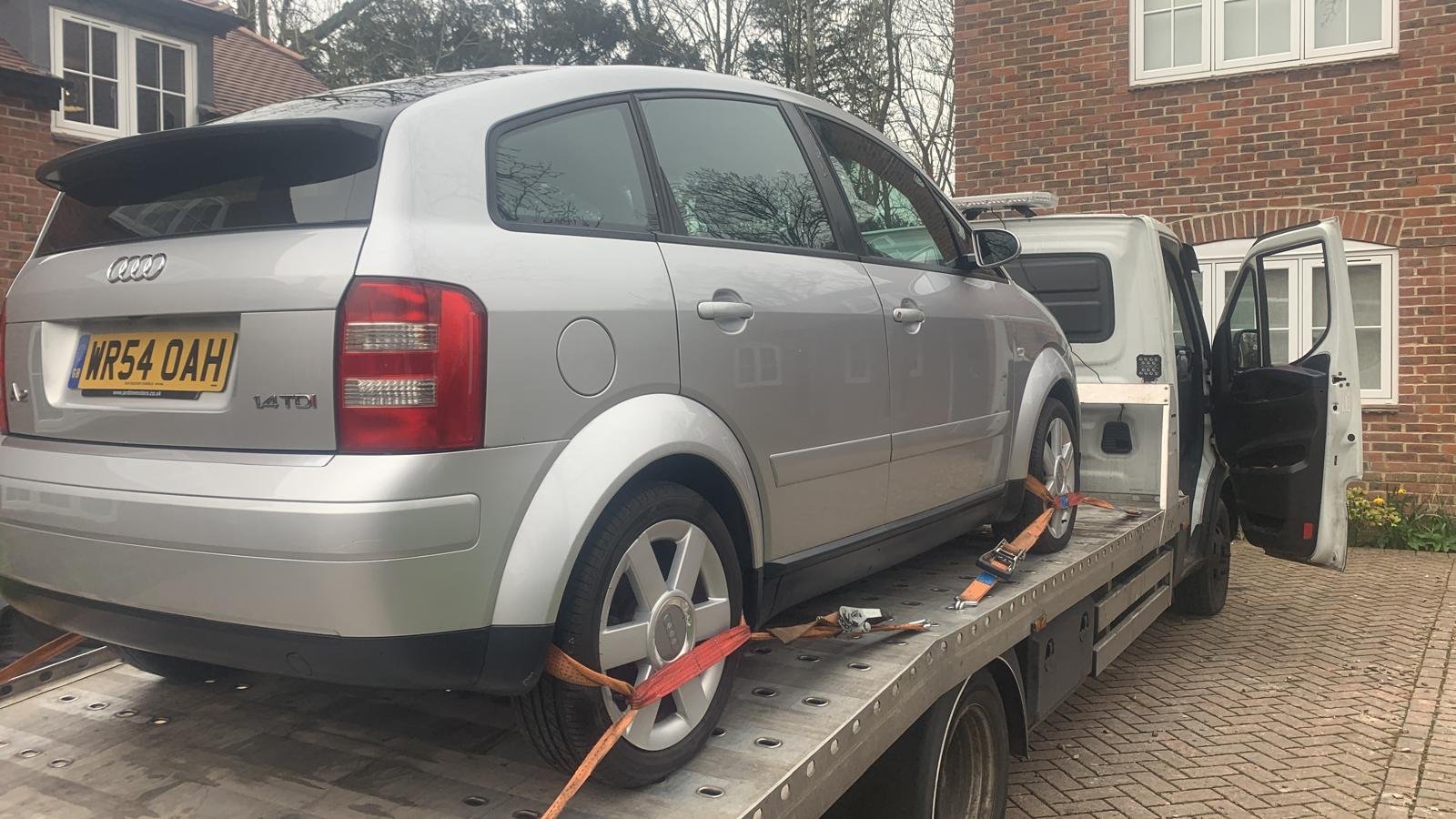 A silver Audi A3 car on a flatbed tow truck, secured with straps, parked on a brick driveway in front of a brick house.