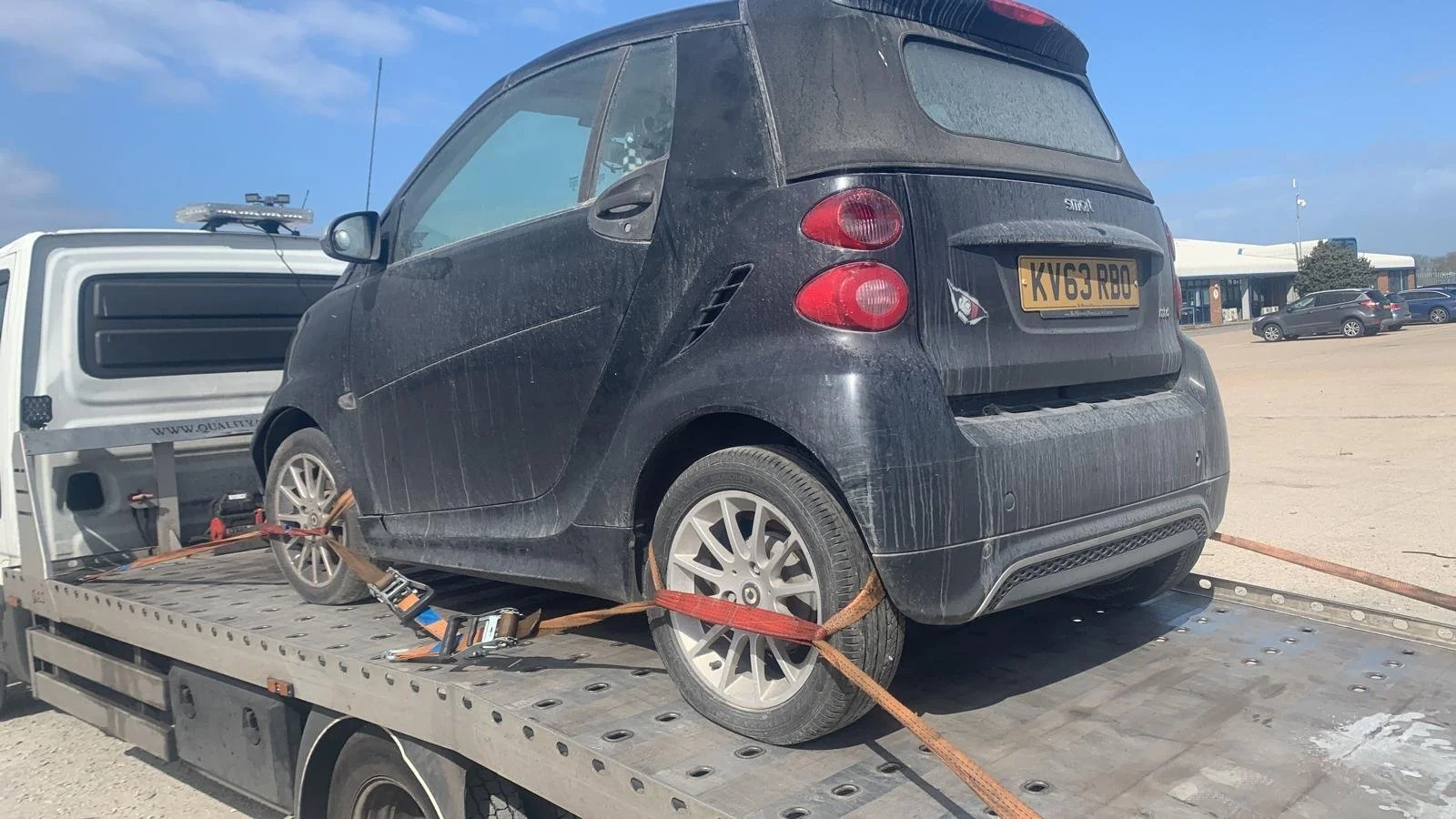 A black Smart car secured with straps on a flatbed tow truck, with a parking lot and building in the background.