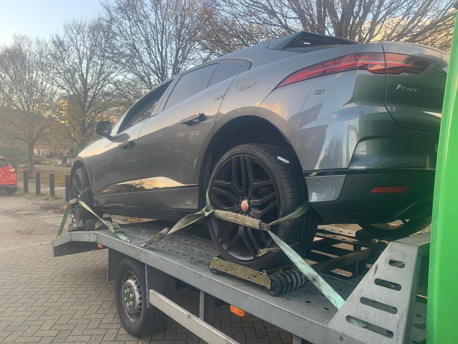 A silver Jaguar I-PACE electric vehicle secured with straps on a flatbed tow truck in an outdoor parking area with leafless trees in the background.