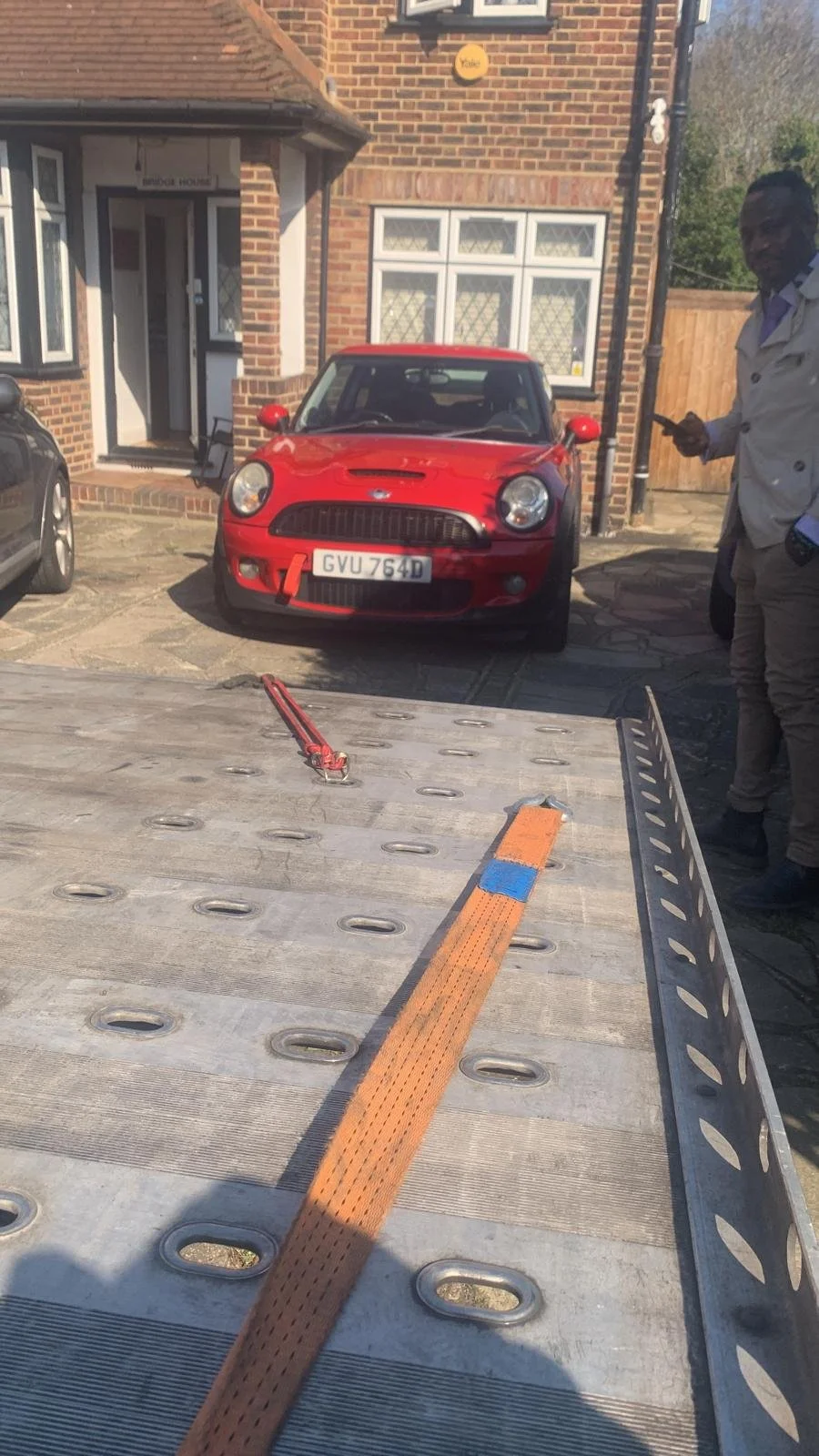 A red Mini Cooper car parked in front of a brick house, with a man standing to the right holding a phone, around a flatbed trailer with orange straps and a tool on it.