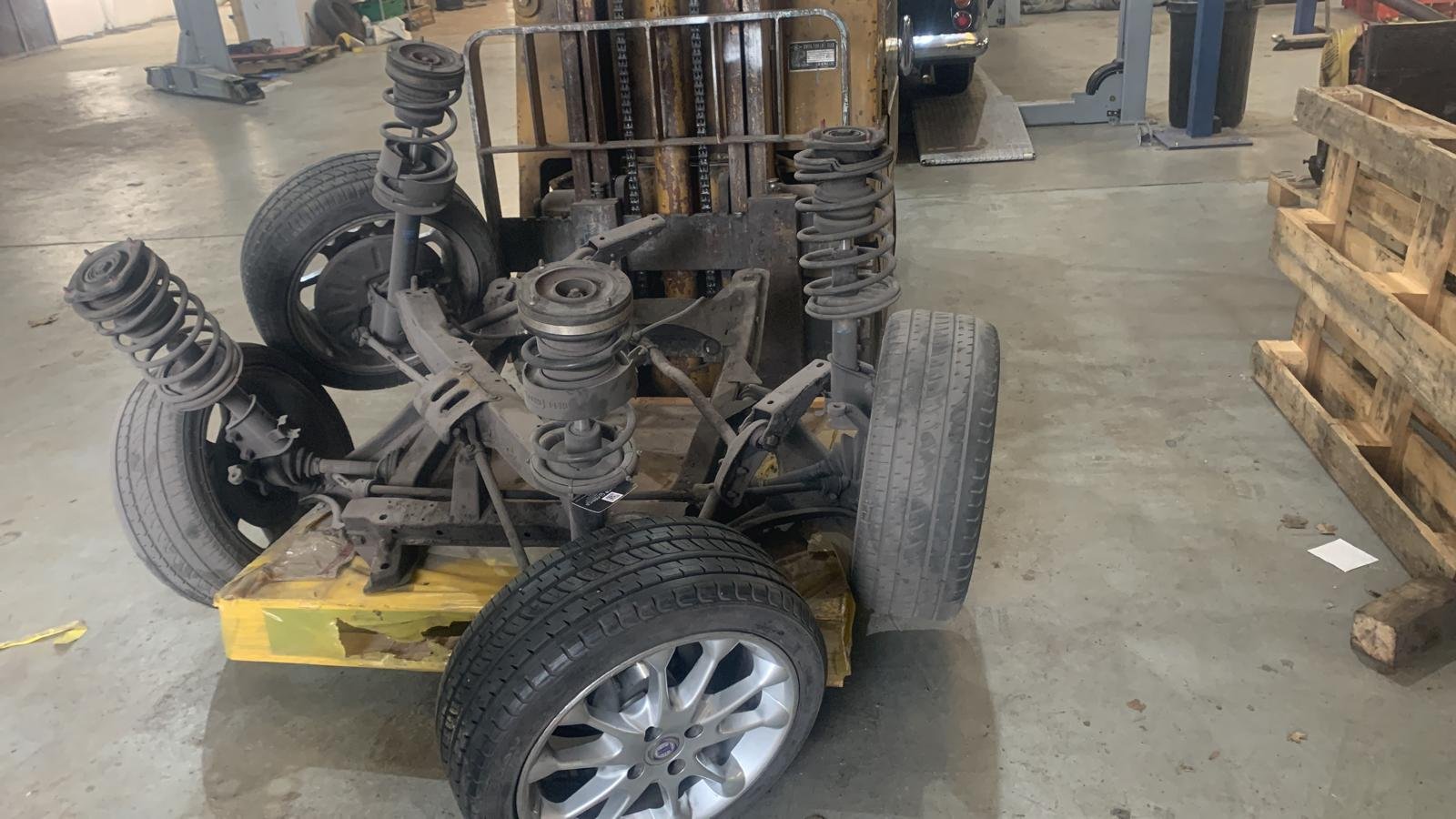 Car chassis with wheels and suspension components in a workshop. The frame is mounted on a yellow base, with tires attached to the wheels and visible steel springs.