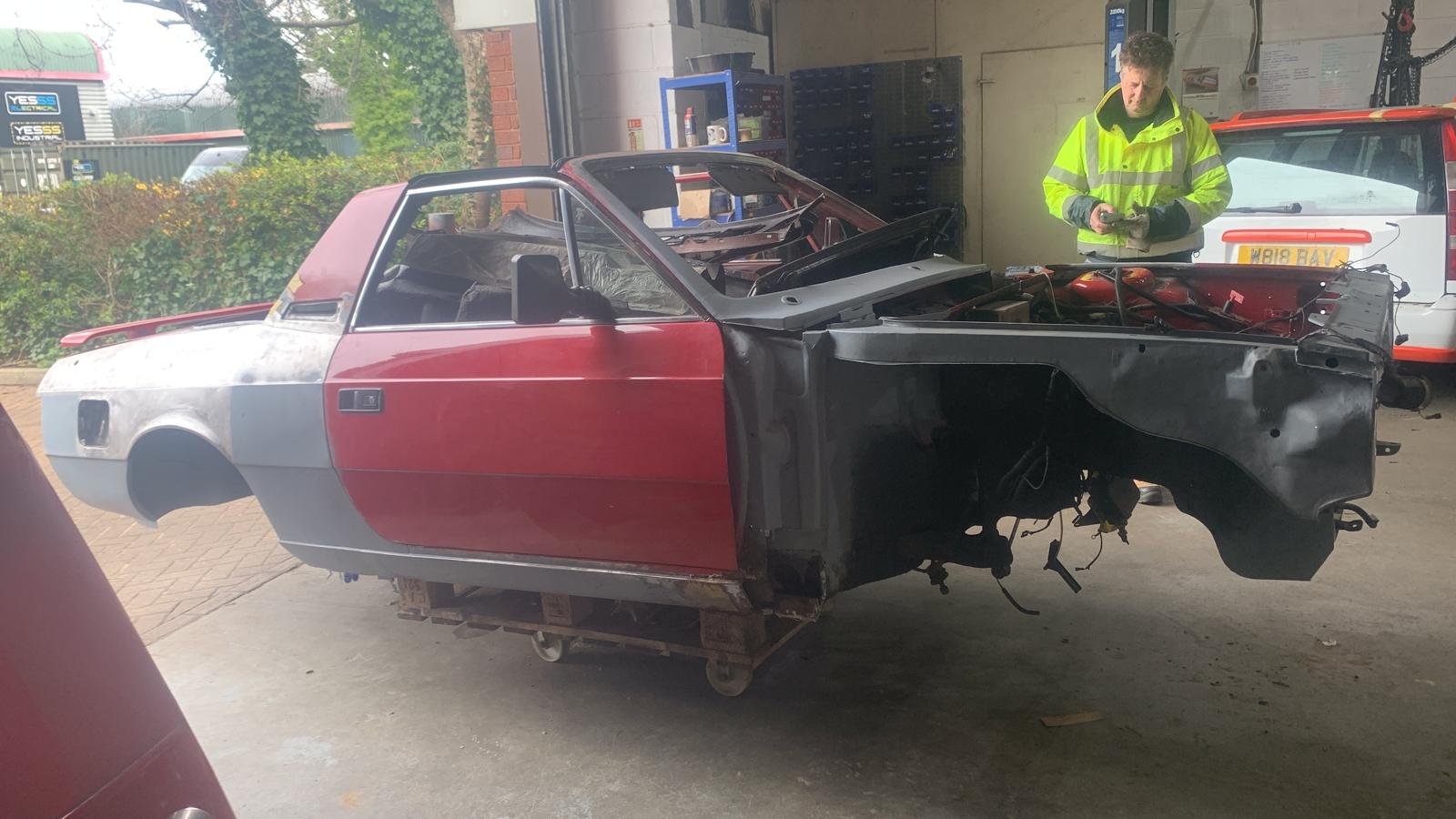 A partially disassembled car with no front wheels in a workshop, with a man in a yellow jacket inspecting the engine area.