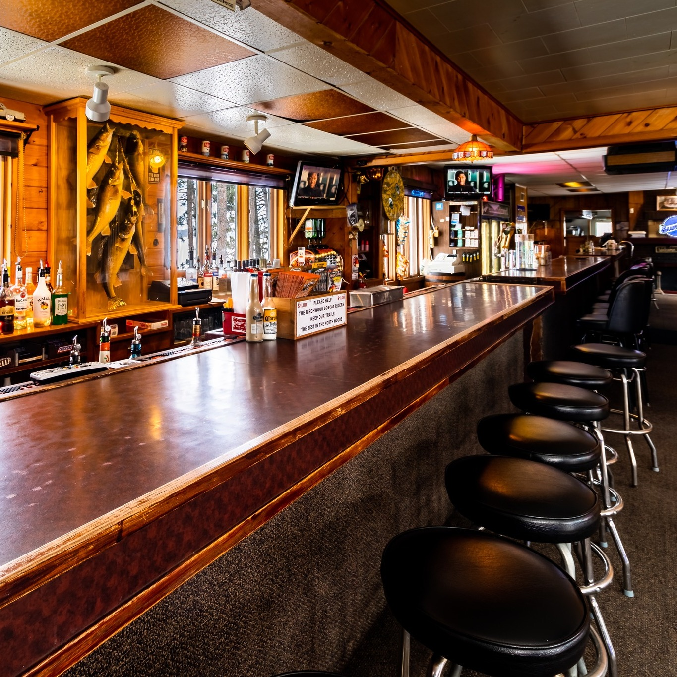 A wooden bar with black barstools, bottles behind the bar, two small TVs on the wall showing a football game, and a window with trees outside in a cozy, rustic pub.