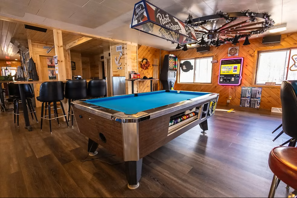 A cozy bar or rec room with a pool table, high chairs, and arcade games, featuring wood paneling and warm lighting.