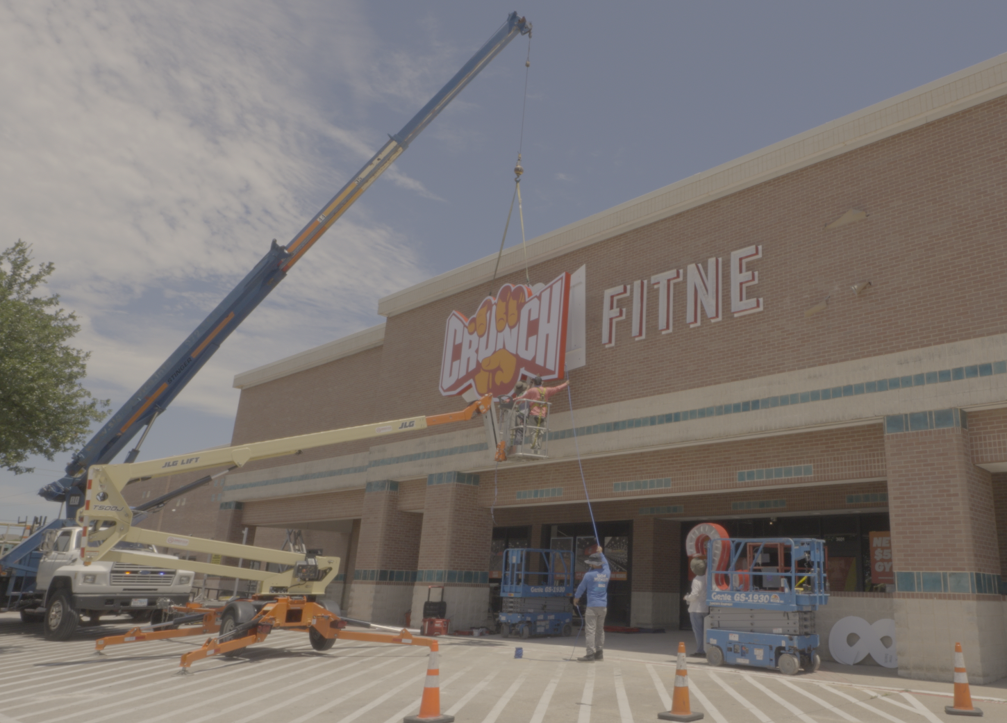 Workers are installing a sign that reads 'Crunch Fitness' on the front of a brick building using a crane and lift, with cones marking the area.