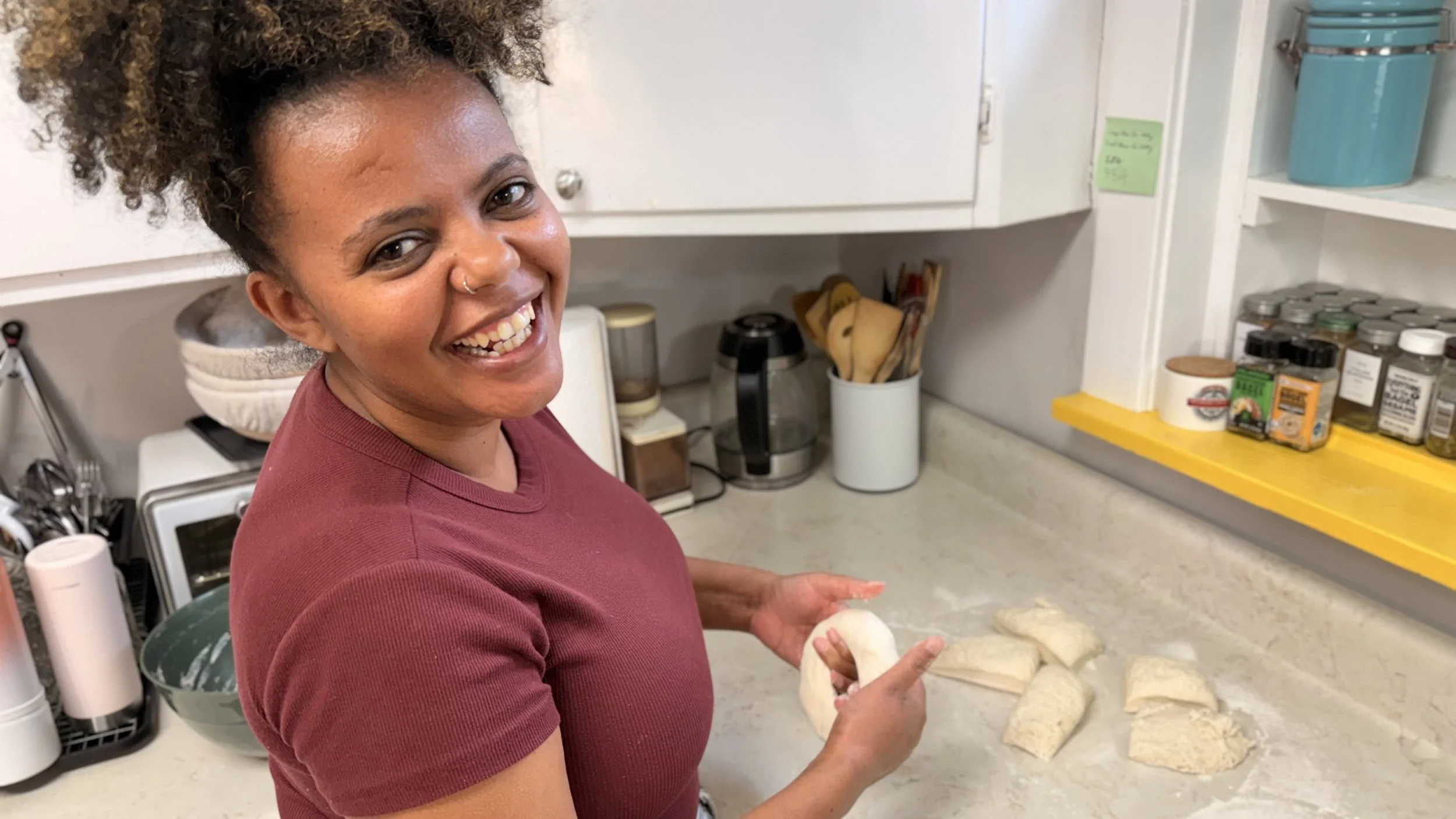 An Ethiopian woman with curly hair and a nose ring smiling in a kitchen, preparing dough on a countertop surrounded by kitchen appliances and pantry items.