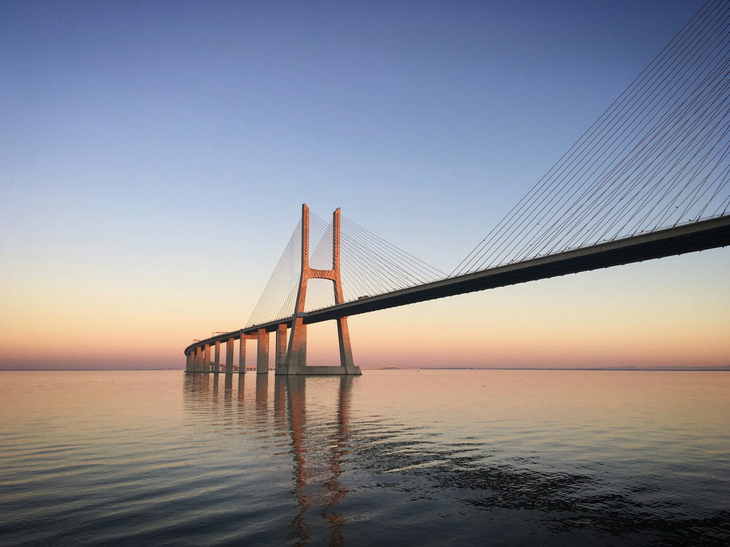 A suspension bridge with tall orange towers and numerous cables spanning over a calm body of water during sunset or sunrise, with a clear sky overhead.