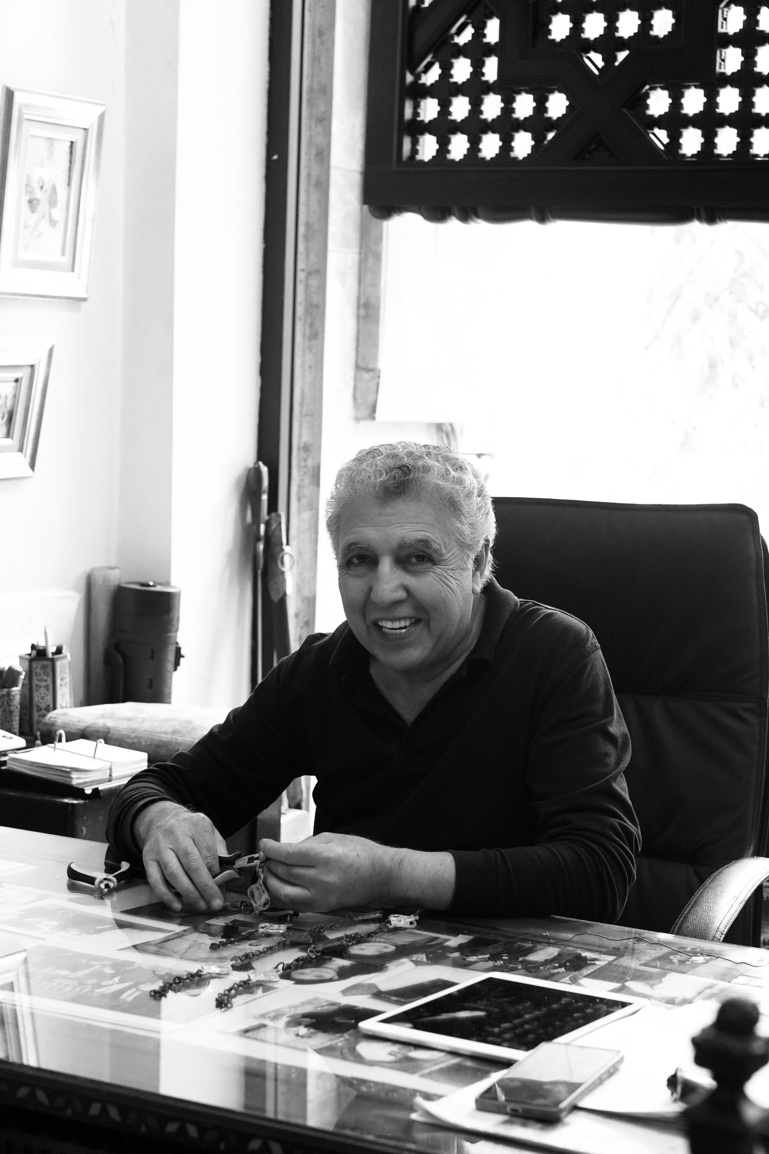 Un homme souriant assis à une table, examinant ou manipulant des objets ou des bijoux, entouré d'outils ou accessoires, dans un espace intérieur avec des œuvres d'art encadrées au mur et un mobilier.