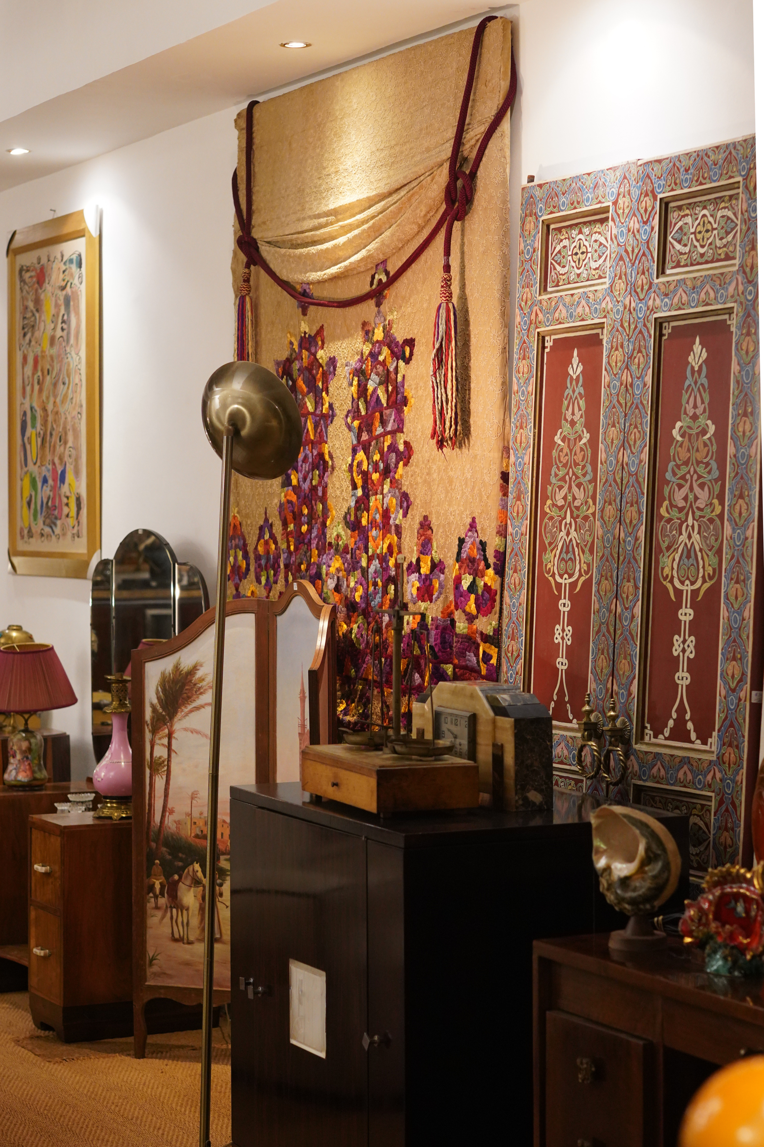 Une pièce décorée avec plusieurs tableaux et meubles anciens, notamment une petite commode en bois, une cabine de télévision noire avec un objet en marbre dessus, et une étagère contenant des objets décoratifs. On aperçoit également un tapis beige et un luminaire en forme de globe en métal.