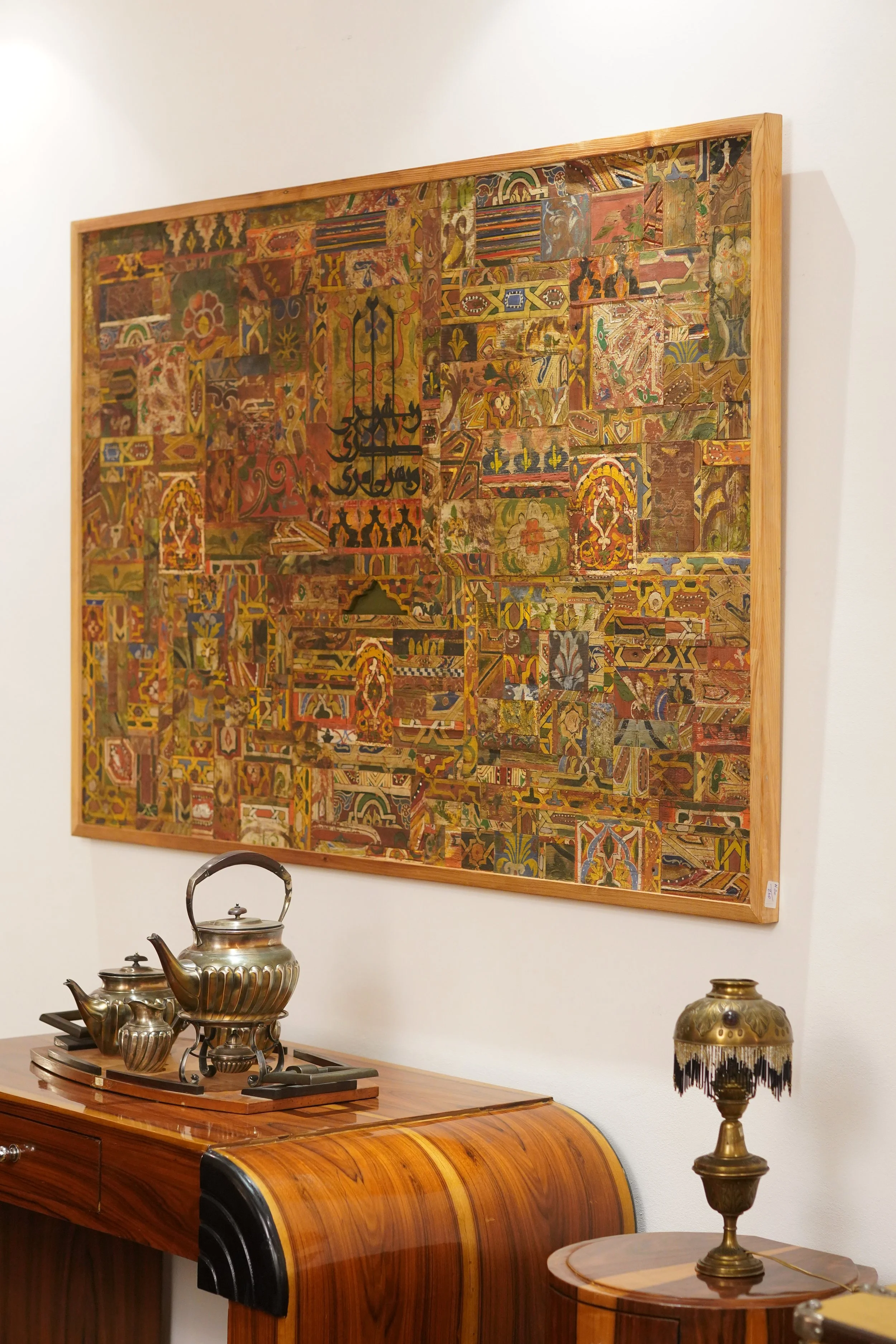 Une œuvre d'art murale composée de plusieurs pièces colorées, encadrée en bois, suspendue au mur. En dessous, une table en bois accueille un service à thé en métal argenté et une lampe ancienne en laiton avec un abat-jour de style marocain.