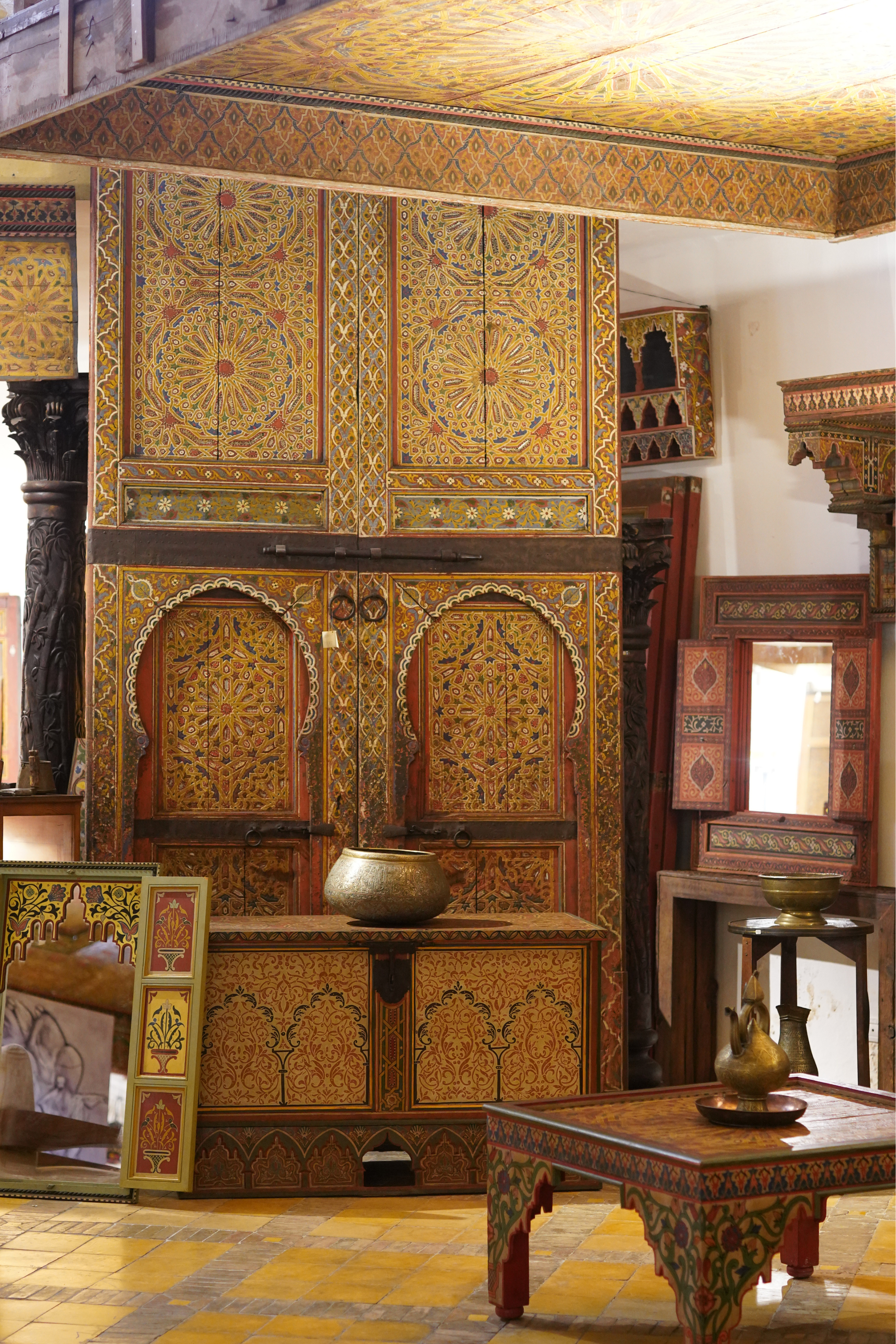 Une pièce décorée avec des meubles peints à la main avec un motif géométrique et floral coloré, comprenant une grande armoire, une table, et plusieurs petites étagères. Objets en métal jaune, comme un vase, sont placés sur certains meubles.Couleurs principalement jaunes, rouges, et noires.