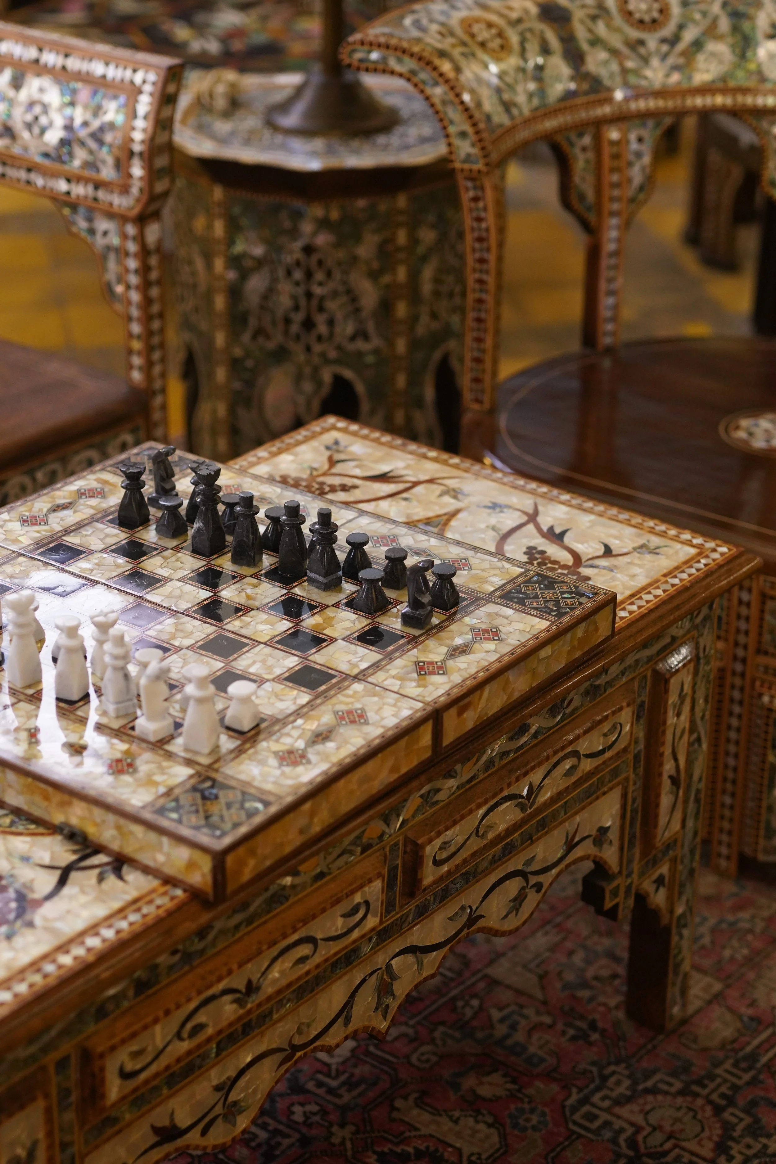 Un jeu d'échecs en bois noir et blanc placé sur une table en marqueterie décorative, dans une pièce ornée de meubles en bois finement incrustés.