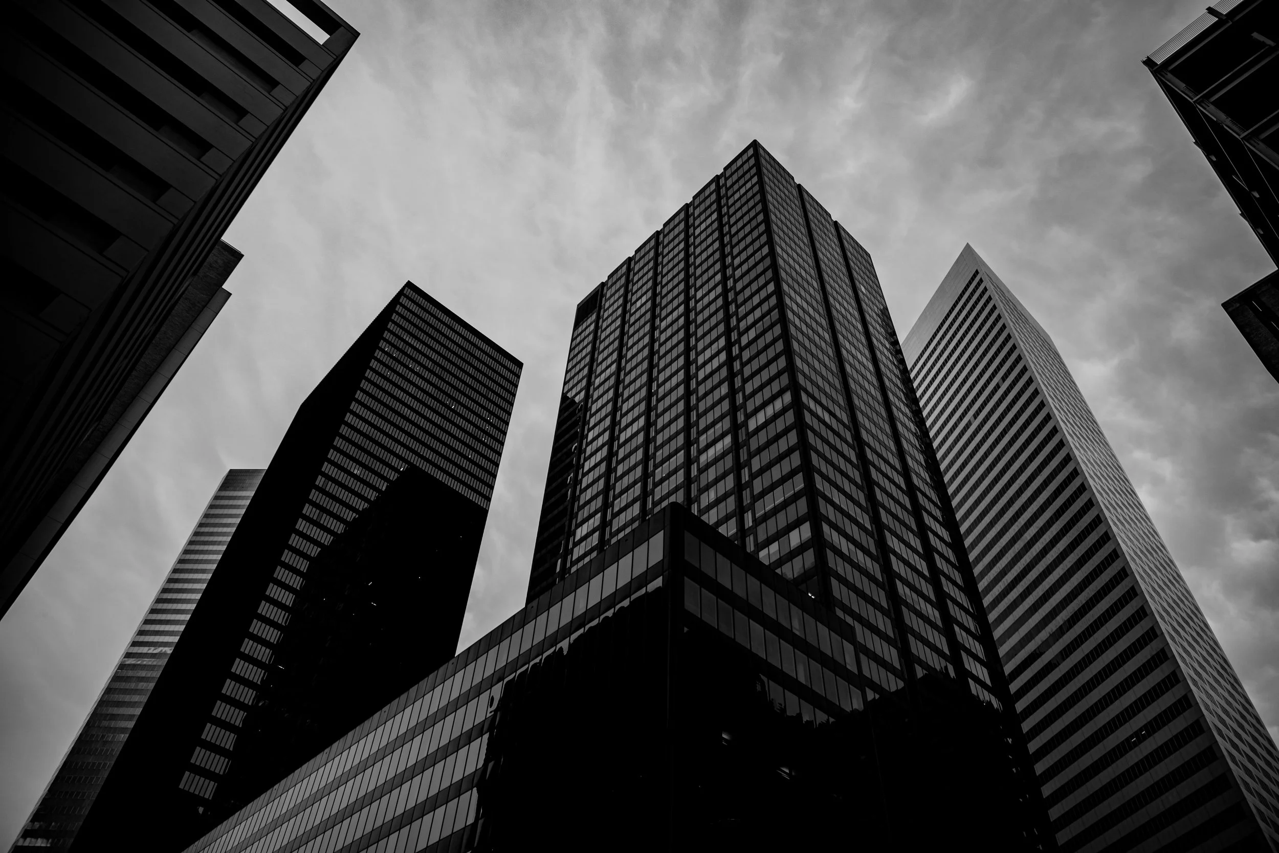 Black and white photograph of tall office buildings viewed from below, with a cloudy sky in the background.