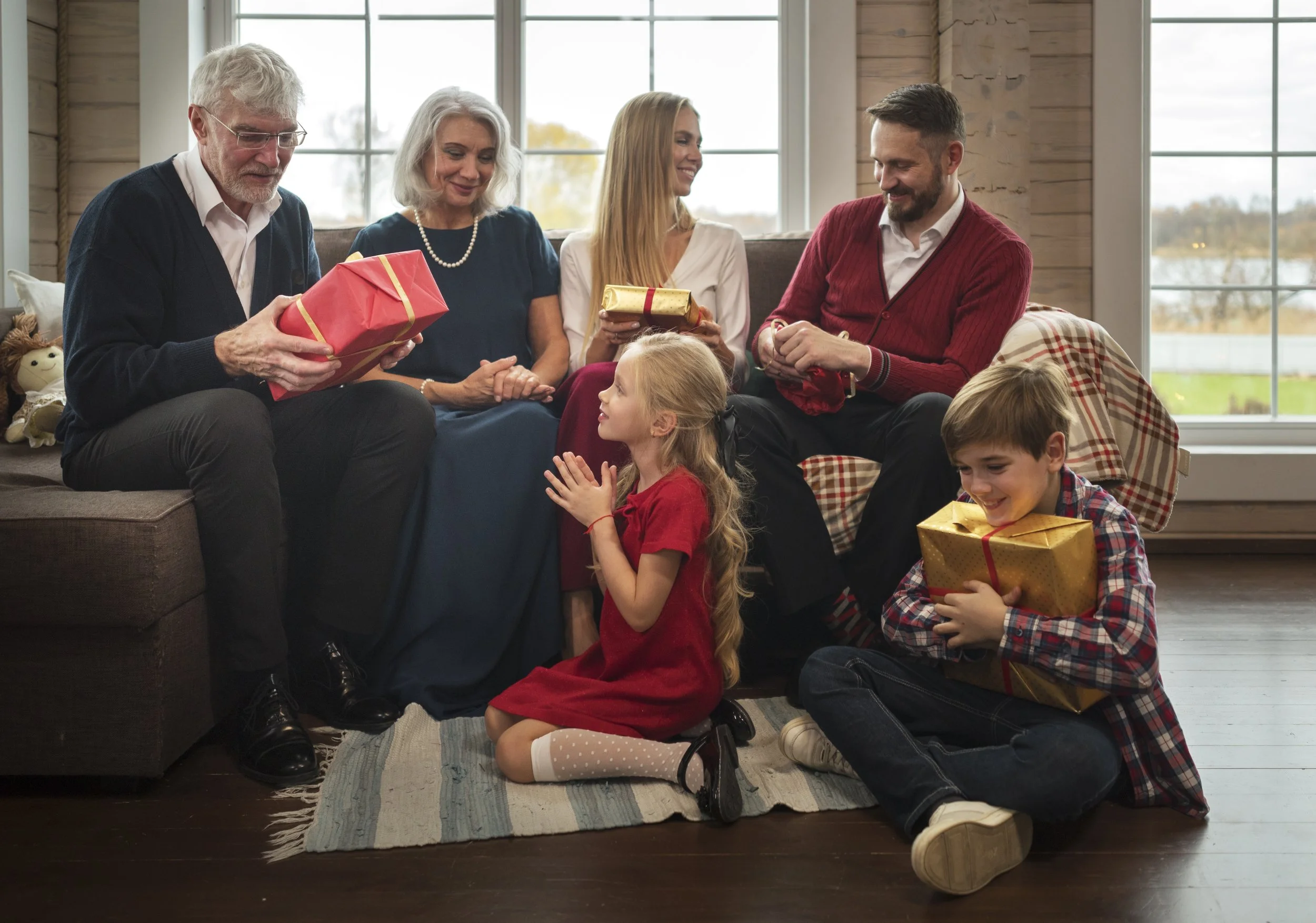 A multigenerational family opening presents in a cozy living room with large windows, wood-paneled walls, and a colorful rug. The group includes elderly, middle-aged, and young children, all smiling and engaged in the gift-opening celebration.