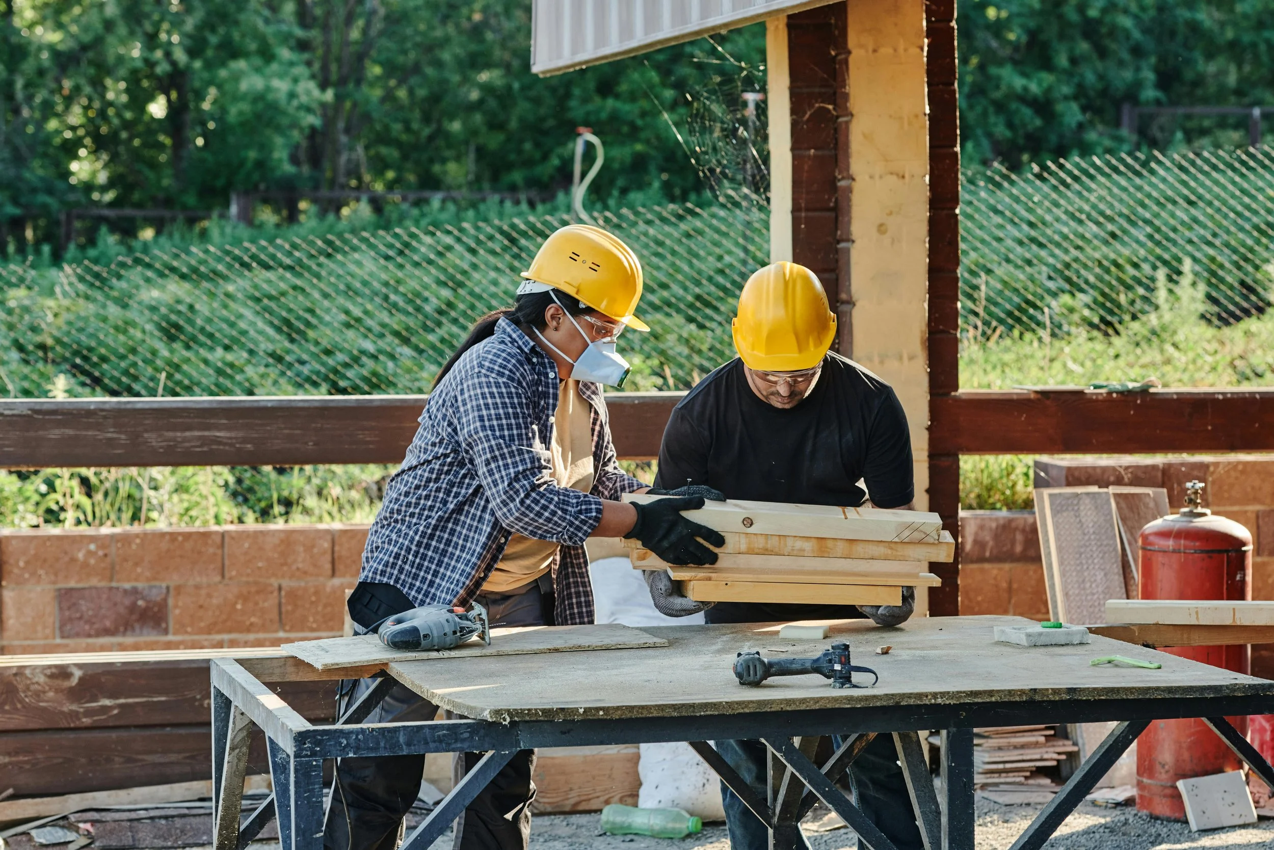 Two construction workers wearing yellow safety helmets, face masks, and gloves working on a woodworking project outdoors during daytime. They are focused on a wooden piece on a work table, with tools like drills nearby. There is a brick wall and a green fence in the background.