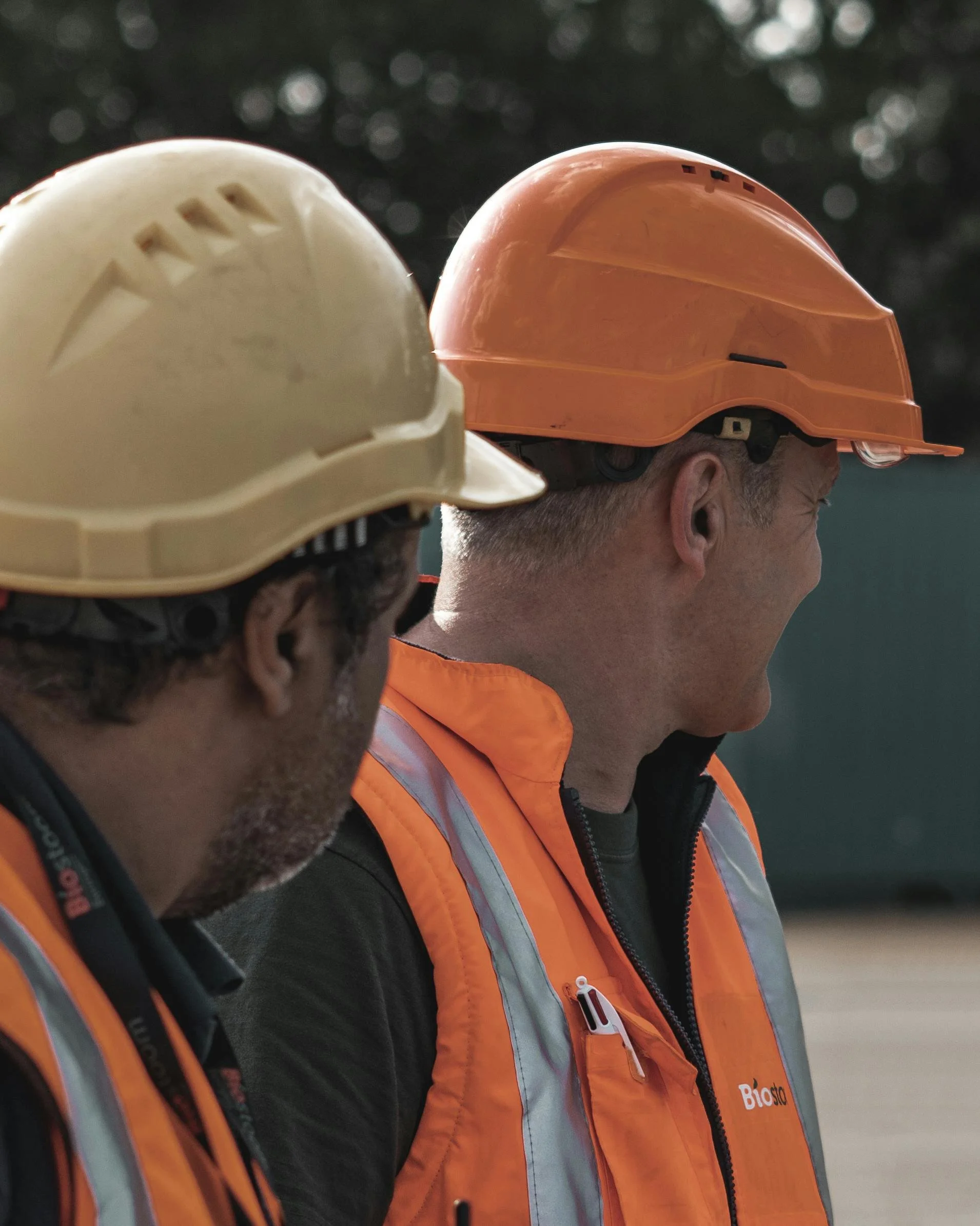 Two construction workers wearing orange safety vests and helmets, standing outdoors against a blurry background.