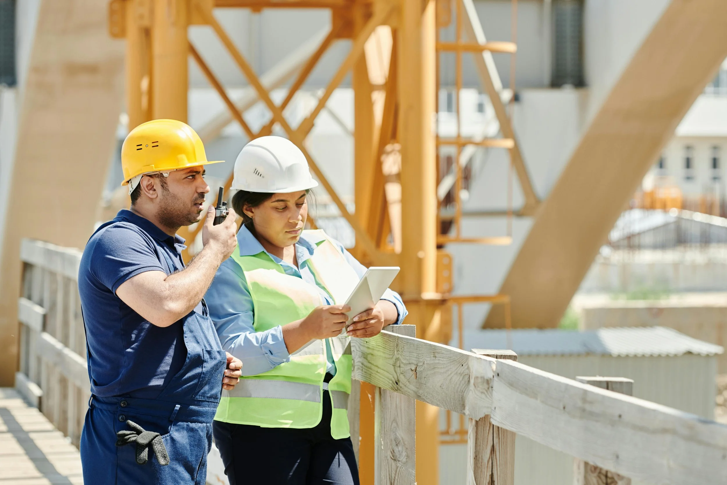 Two construction workers, one man and one woman, wearing safety helmets and vests, inspecting a construction site with wooden structures and a crane in the background.
