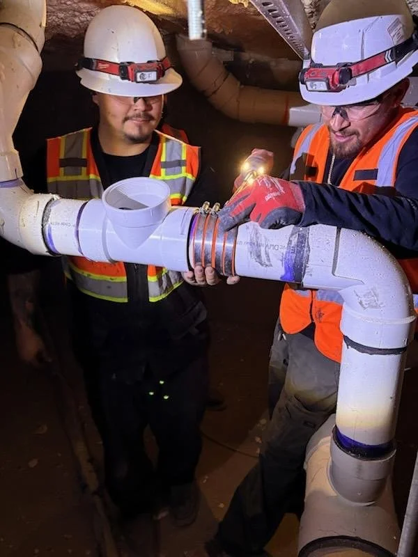 Two A&K Plumbing and Mechanical technicians in safety gear and helmets working together on a large pipeline installation underground.