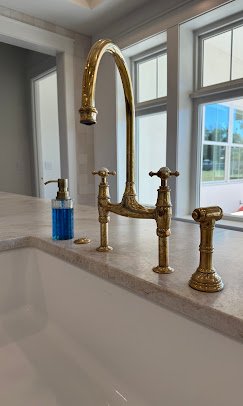 Gold kitchen faucet with two handles on a white countertop in front of a window. Plumbing, HVAC, and mechanical outline icon for A&K Plumbing and Mechanical.