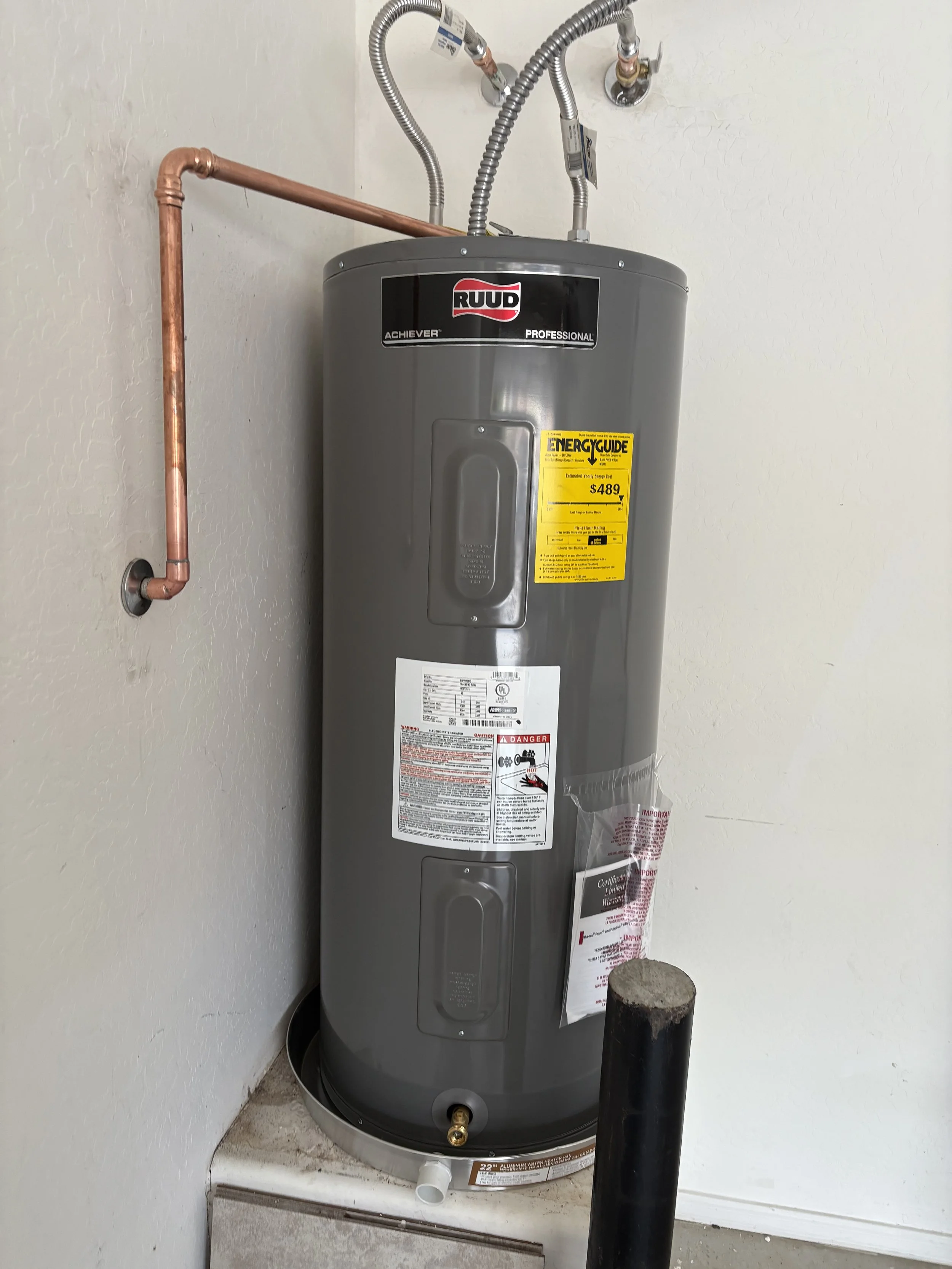 A gray RUUD water heater installed against a white wall with copper and flexible metal pipes connected to it, and a black pipe on the ground nearby. Plumbing, HVAC, and mechanical outline icon for A&K Plumbing and Mechanical.