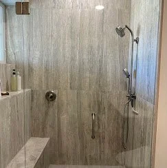 Shower area with beige tiled walls, a handheld showerhead, a fixed showerhead, a small built-in bench, and a grab bar. Plumbing, HVAC, and mechanical outline icon for A&K Plumbing and Mechanical.