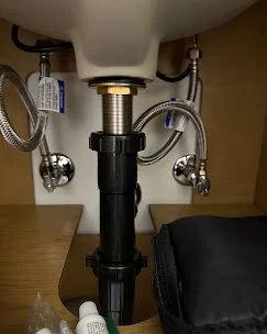 A view of a kitchen sink's plumbing underneath, including pipes, hoses, and a black P-trap, with a wooden cabinet in the background. Plumbing, HVAC, and mechanical outline icon for A&K Plumbing and Mechanical.