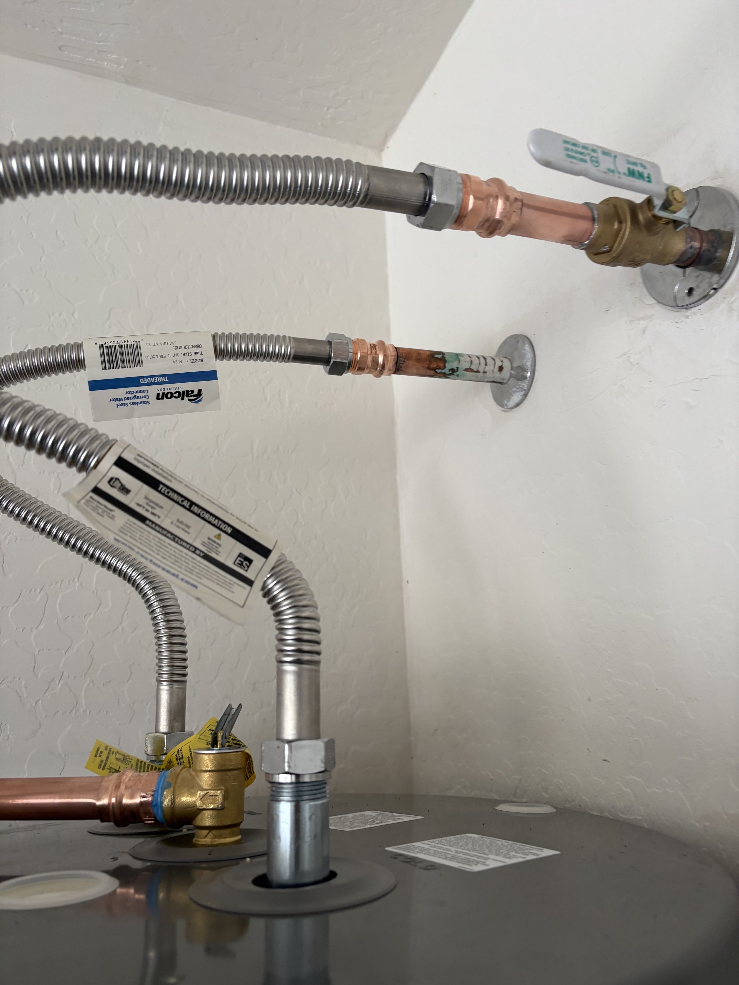 Close-up view of plumbing pipes and fittings, including copper, brass, and flexible metal tubing, connected to a wall and floor in a building's utility area. Plumbing, HVAC, and mechanical outline icon for A&K Plumbing and Mechanical.