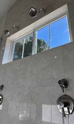 A bathroom with a large horizontal window, three shower heads, and a hand-held shower spray on a gray tiled wall. Plumbing, HVAC, and mechanical outline icon for A&K Plumbing and Mechanical.