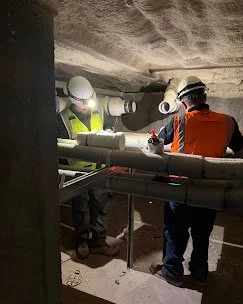 A&K technicians in a tunnel inspecting pipes and wearing safety gear. Plumbing, HVAC, and mechanical outline icon for A&K Plumbing and Mechanical.