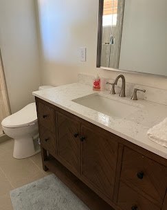 Bathroom with a wooden vanity, marble countertop, and a large mirror. Toilet next to the vanity, and a small blue rug on the floor. Plumbing, HVAC, and mechanical outline icon for A&K Plumbing and Mechanical.