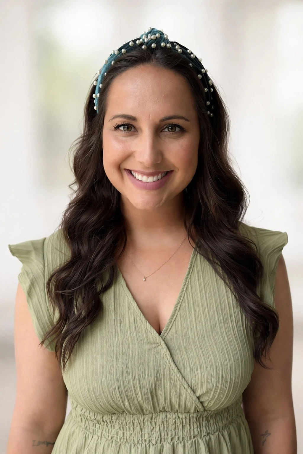 A smiling woman with long dark wavy hair wearing a light green dress with cap sleeves and a pearl headband.