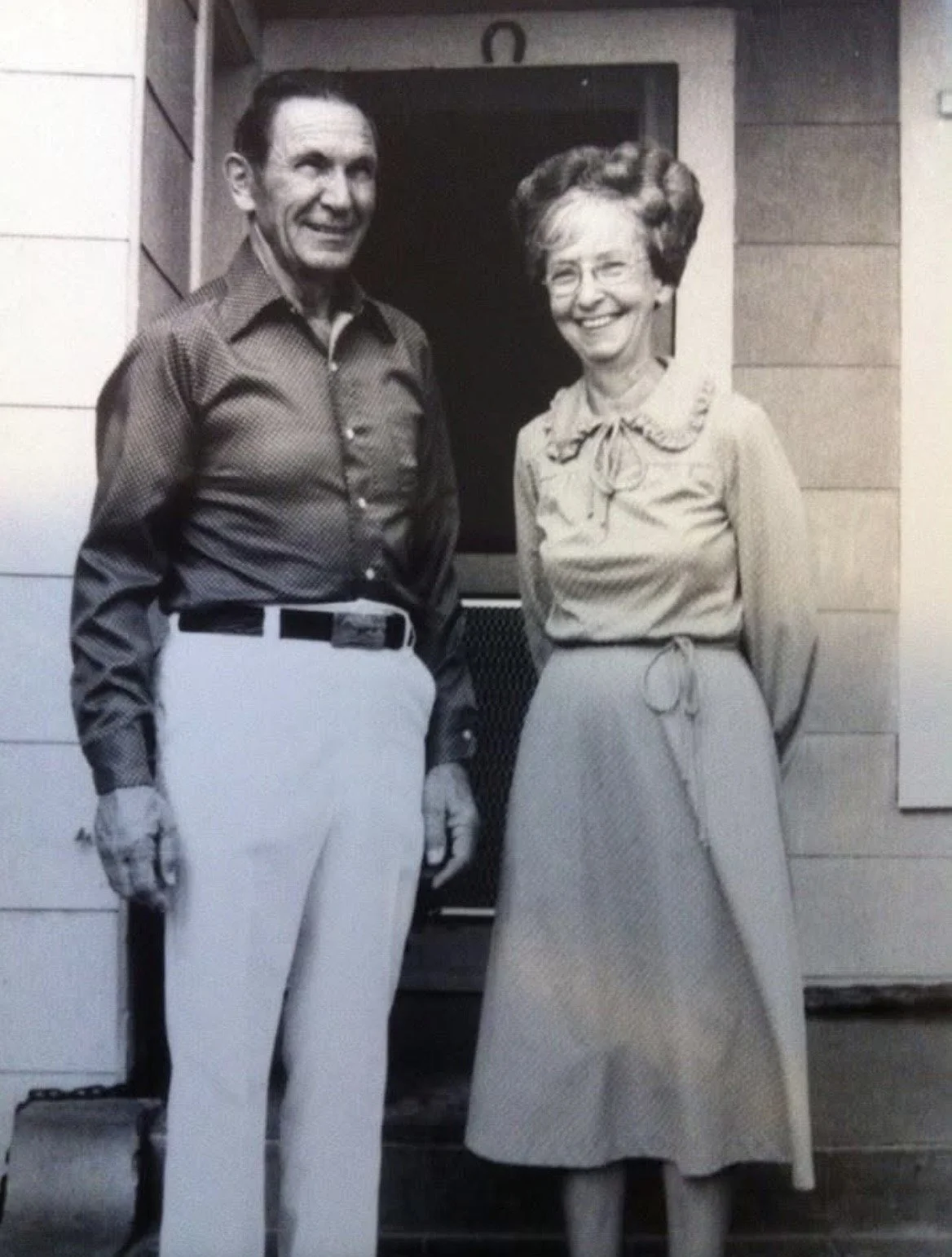 A black-and-white photo of a man and a woman standing side by side outside a building, smiling at the camera. The man is wearing a checkered shirt and white pants, and the woman is wearing a long skirt and a blouse with a bow at the collar.