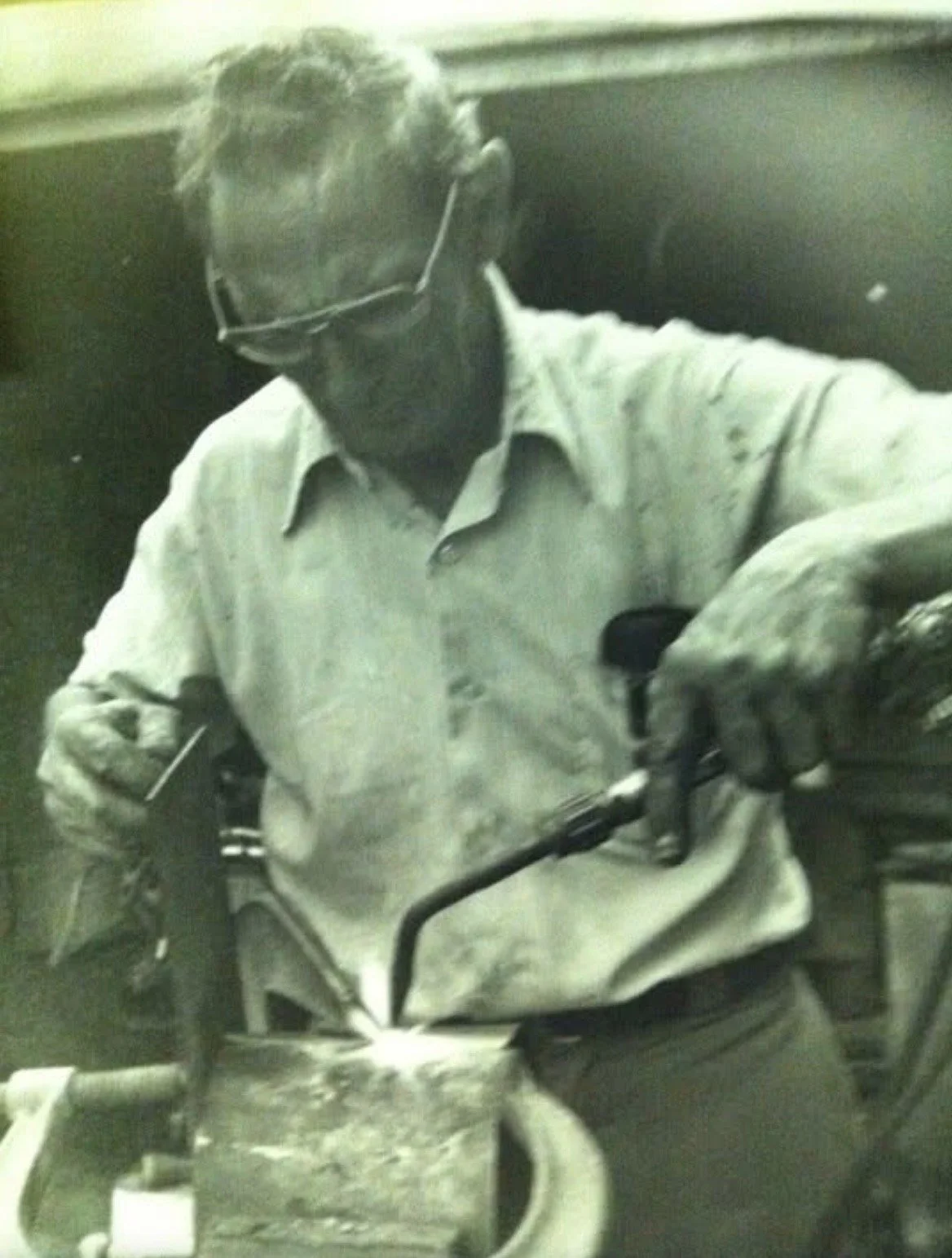 A man wearing glasses and a light-colored shirt working with a blowtorch or similar tool in a workshop or industrial setting.